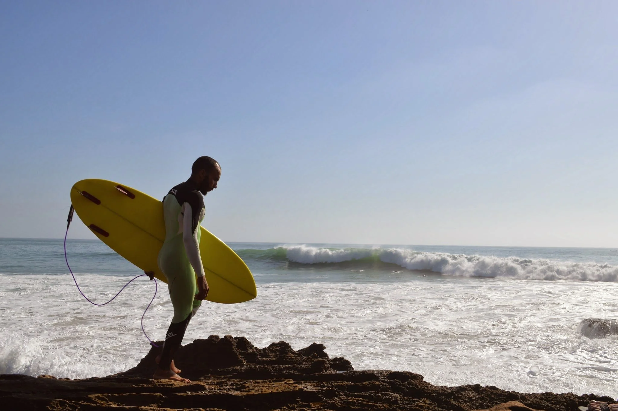 A man in surfing gear holding a yellow surfboard, walking on rocks at the beach with ocean waves in the background under a clear sky.
