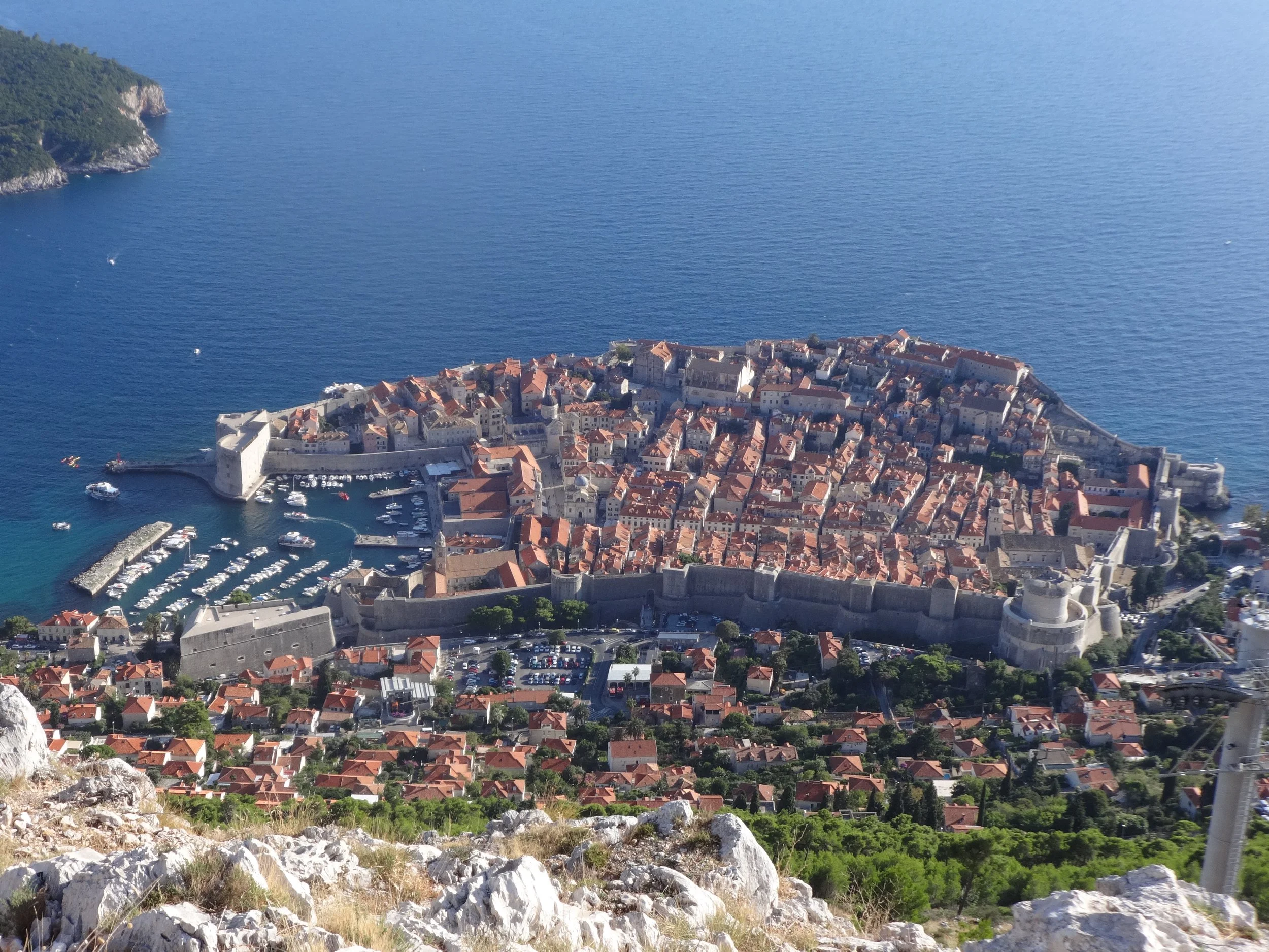 Aerial view of a historic coastal city with stone walls, red-roofed buildings, a marina with boats, and surrounding waters.