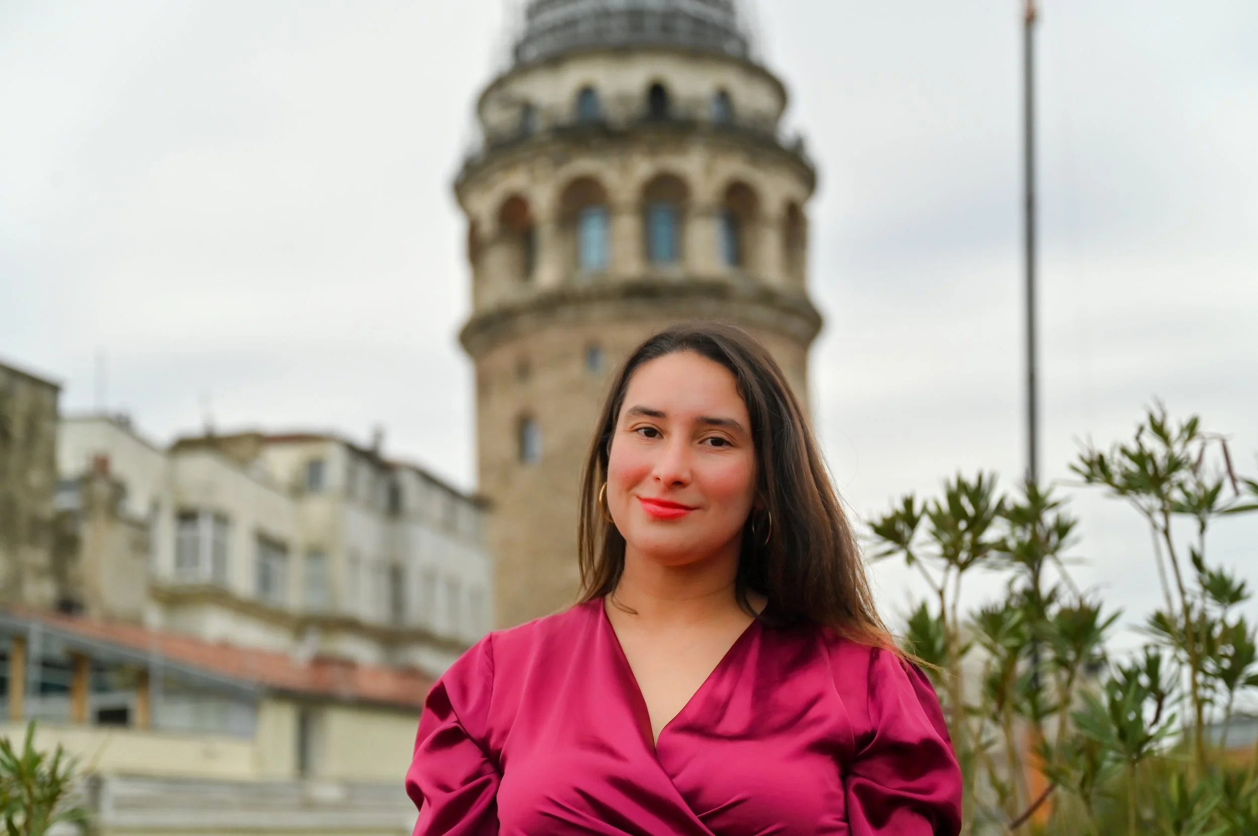 A woman with long brown hair, wearing a pink blouse, standing outdoors with a historic tower and buildings in the background on a cloudy day.