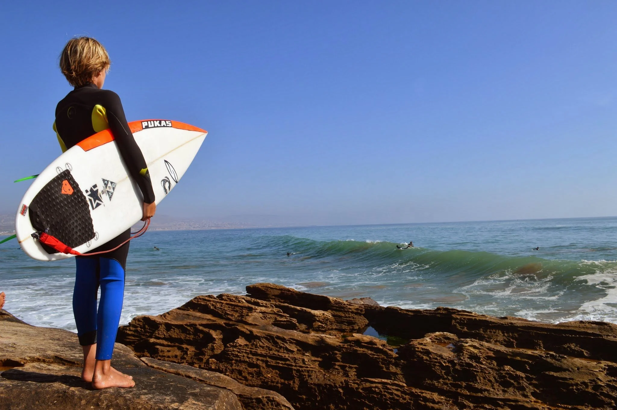 Child standing on rocks at the beach holding a surfboard, looking at the ocean with people surfing in the water.