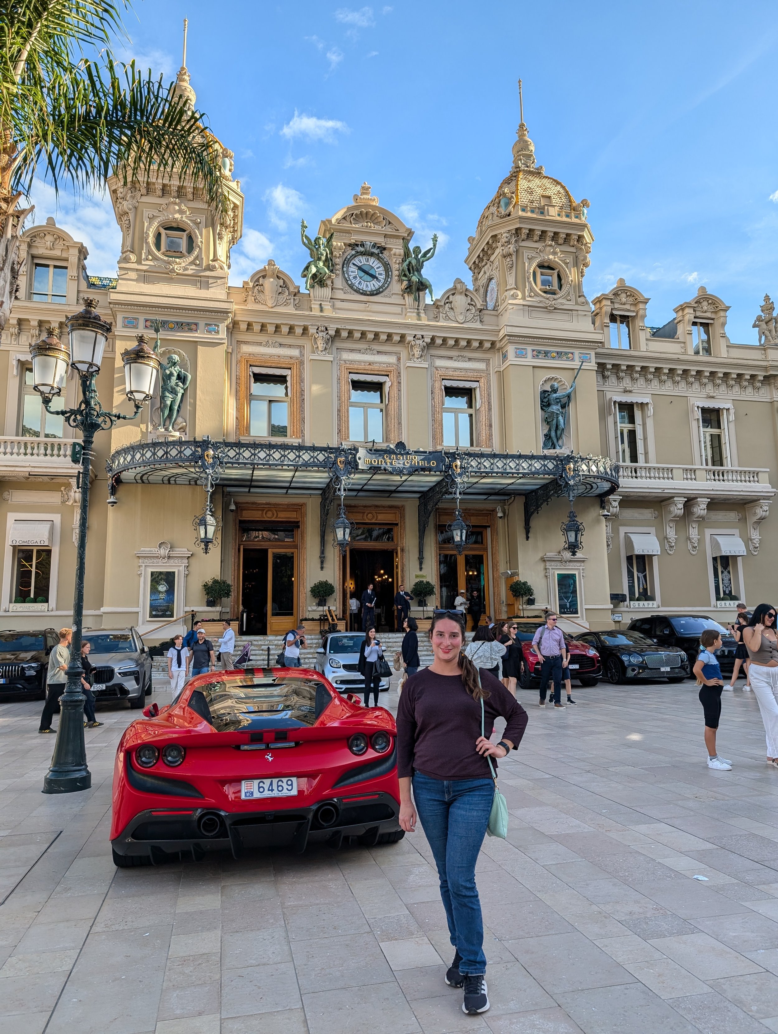 A woman standing in front of a red sports car on a city street with a large ornate building in the background, with people walking around and parked cars nearby.