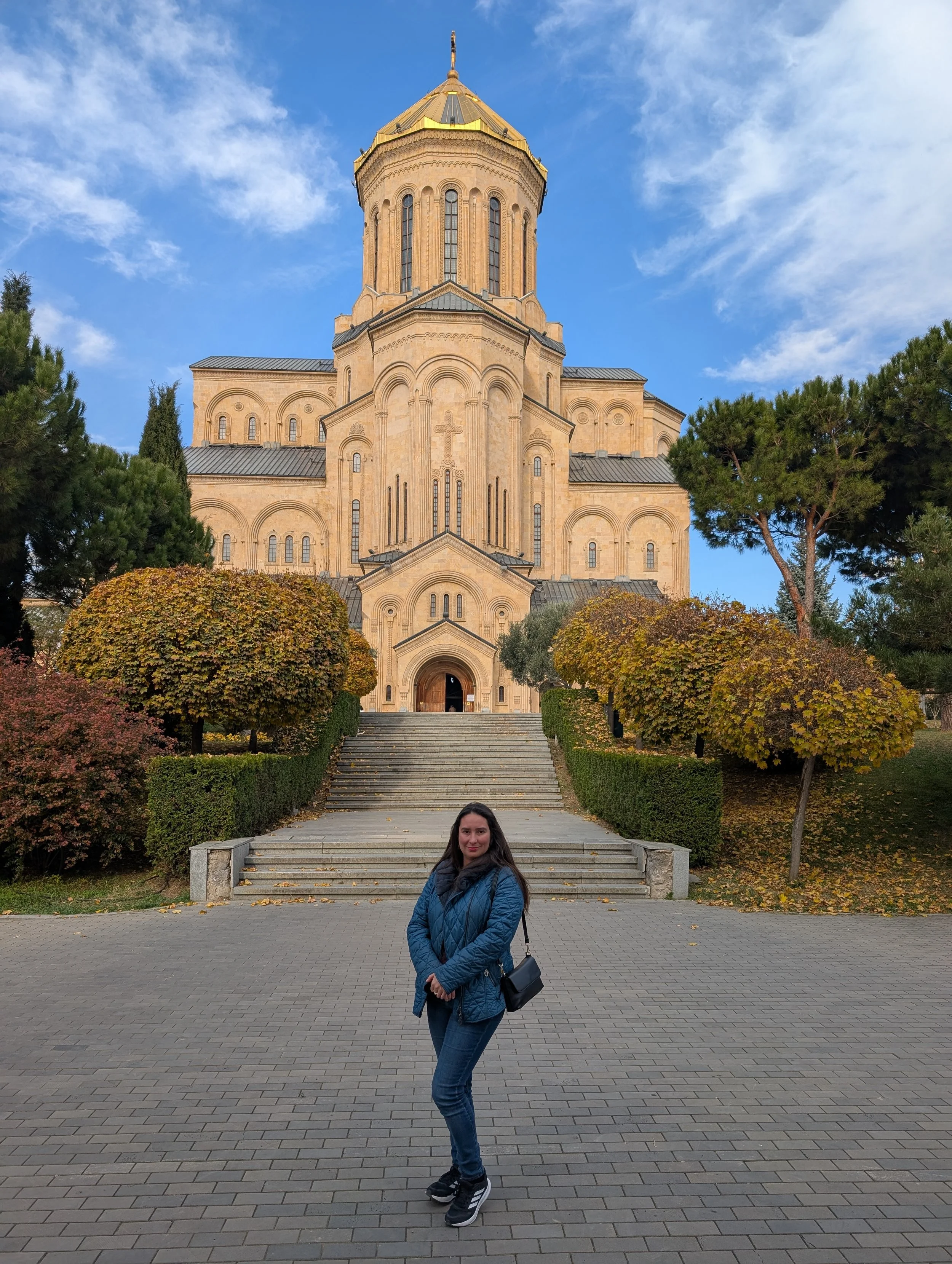 A woman with long dark hair standing on a brick walkway in front of historic church with a large tower, surrounded by trees with fall foliage, under a partly cloudy sky.