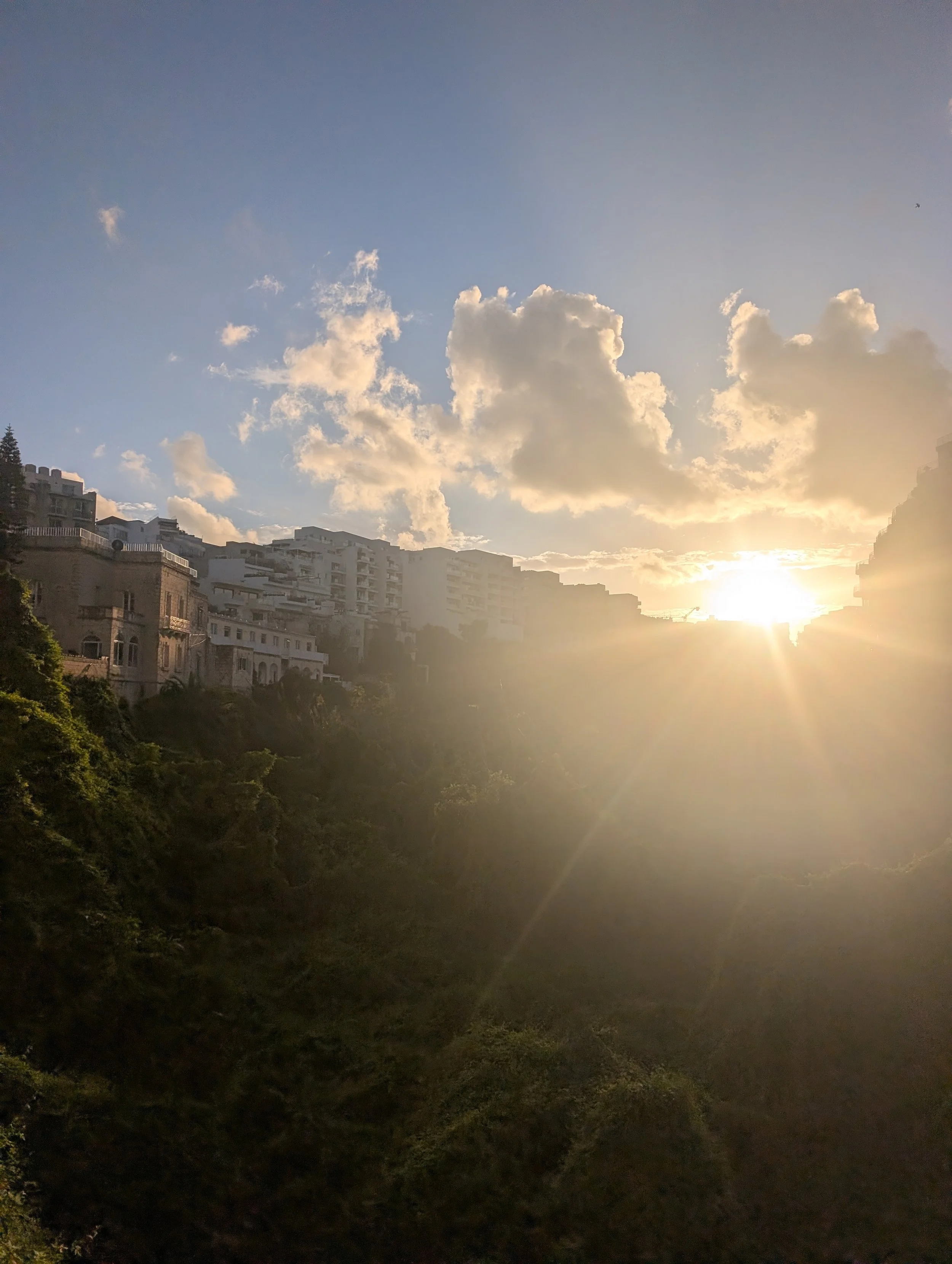 Sunset view over a cityscape with buildings on a hillside, partly cloudy sky, and sunlight streaming through clouds.