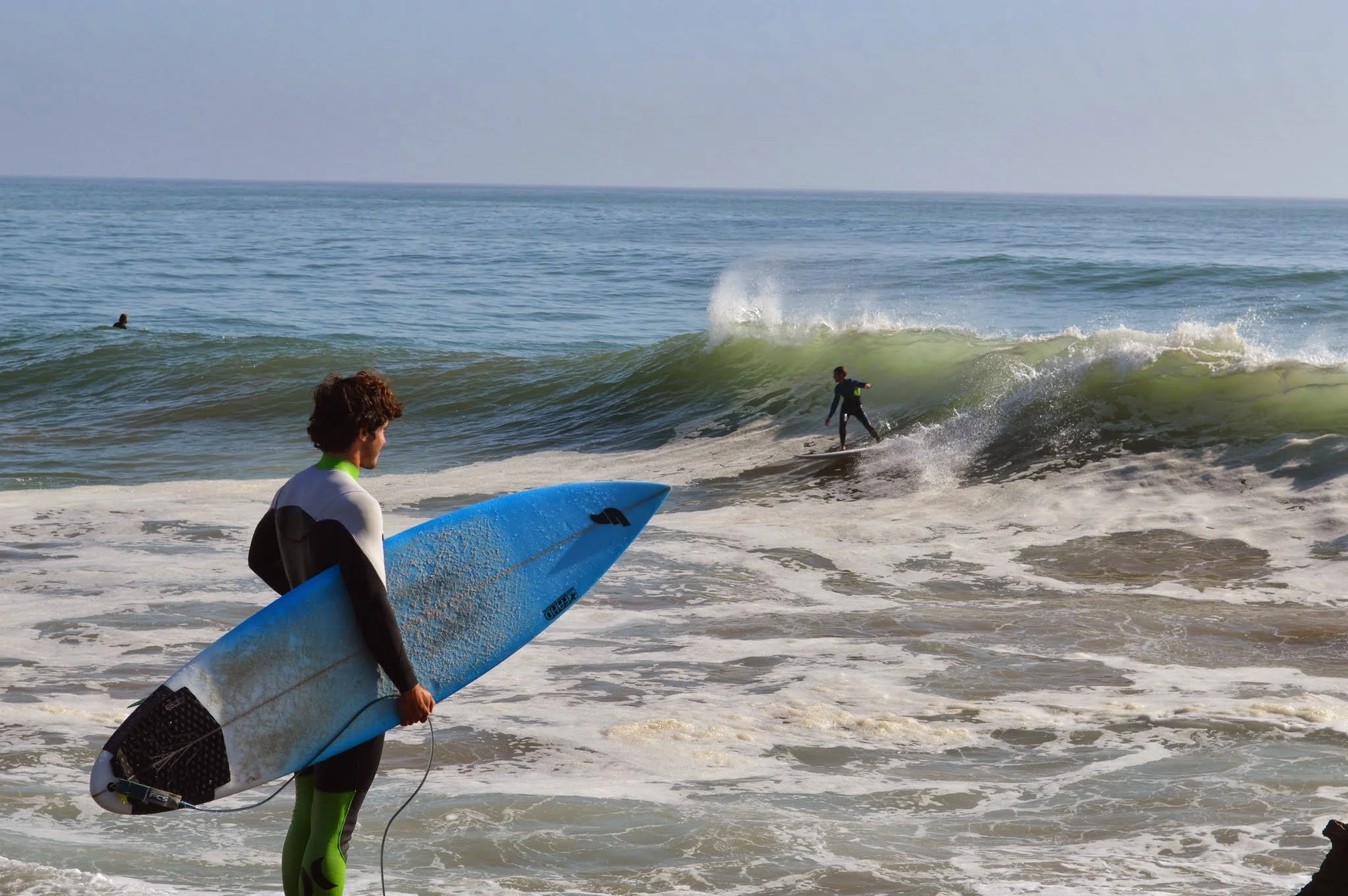 Surfers at the beach, with one preparing to surf and another riding a wave, on a clear day.