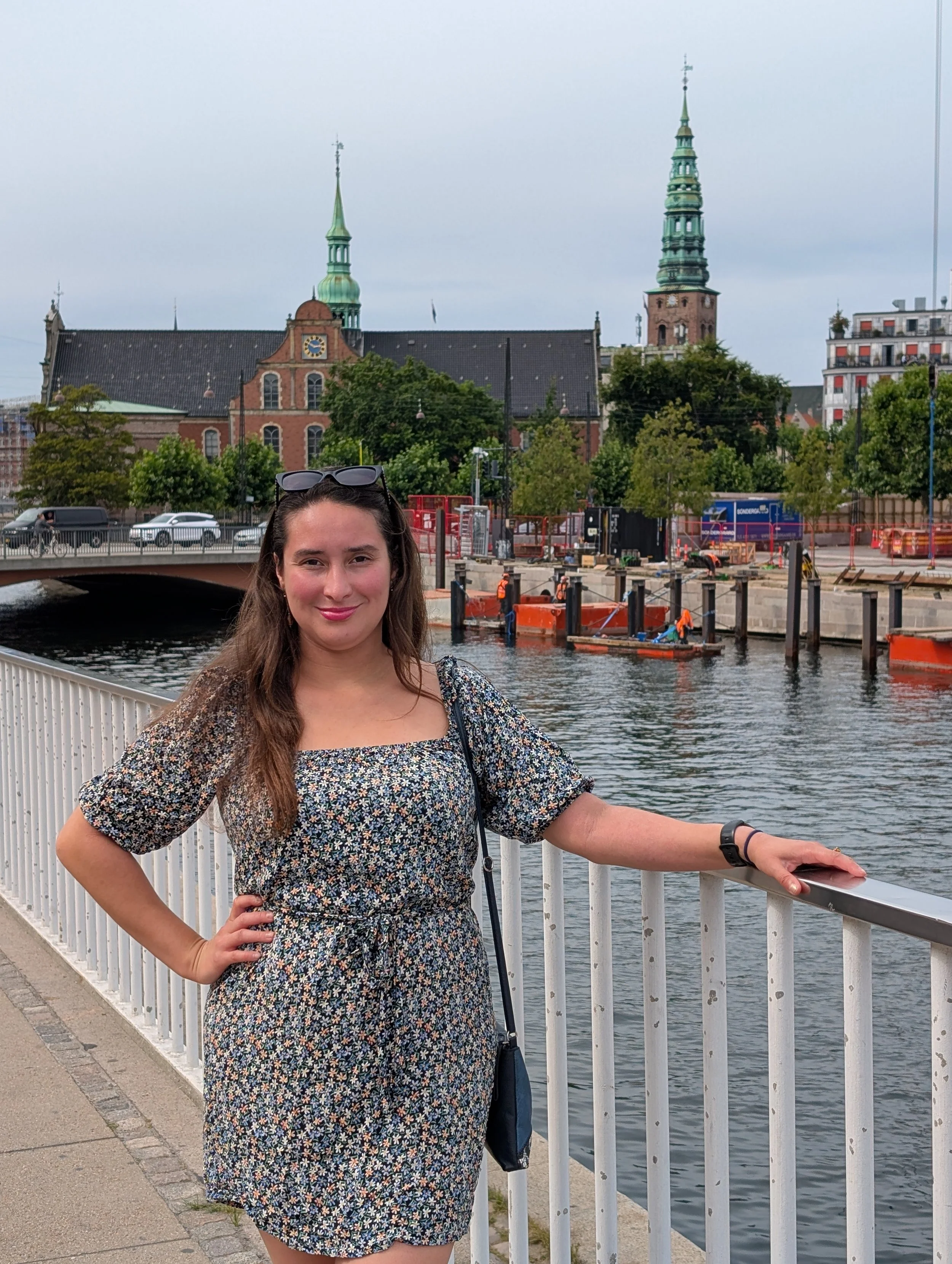 A woman with long brown hair wearing a floral dress standing on a bridge by a river with white railing, with her right hand on her hip and her left hand resting on the railing. There are trees, a church with green spires, and a building in the backgr