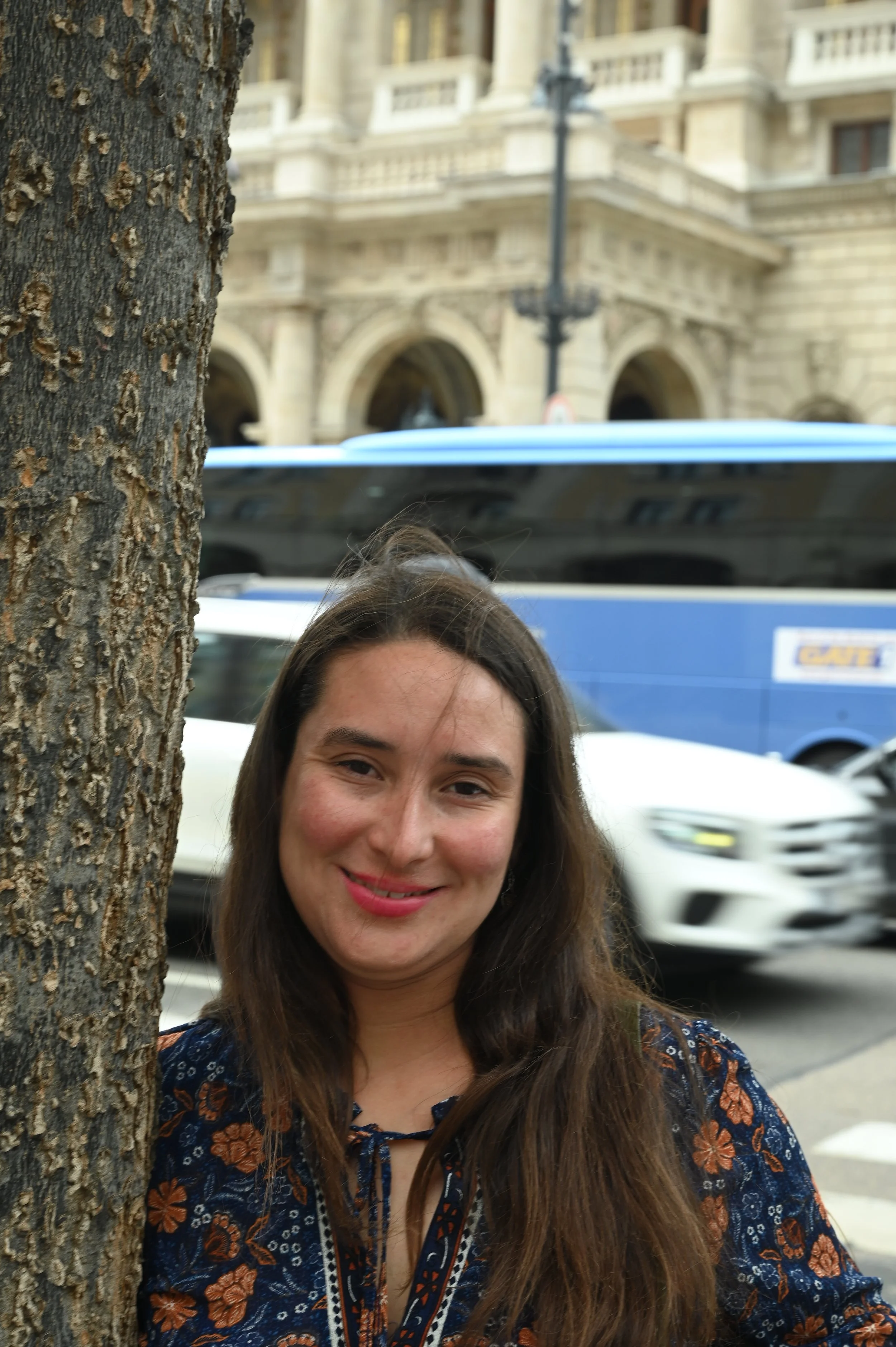 A woman with long brown hair smiling behind a tree on a city street, with a large building and a blue bus in the background.