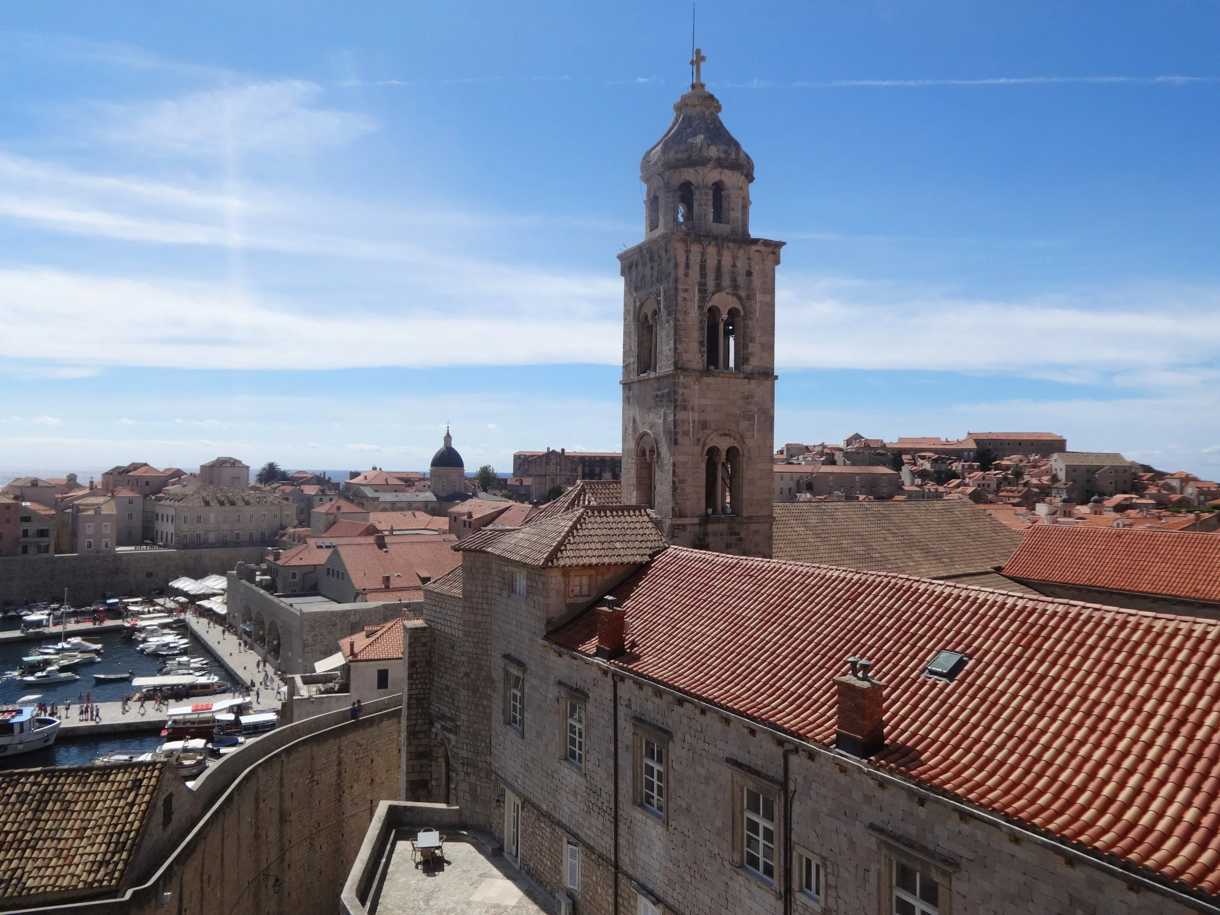 View of a historic cityscape with a tall stone bell tower topped by a cross, red-tiled rooftops, a harbor filled with boats, and a clear blue sky.