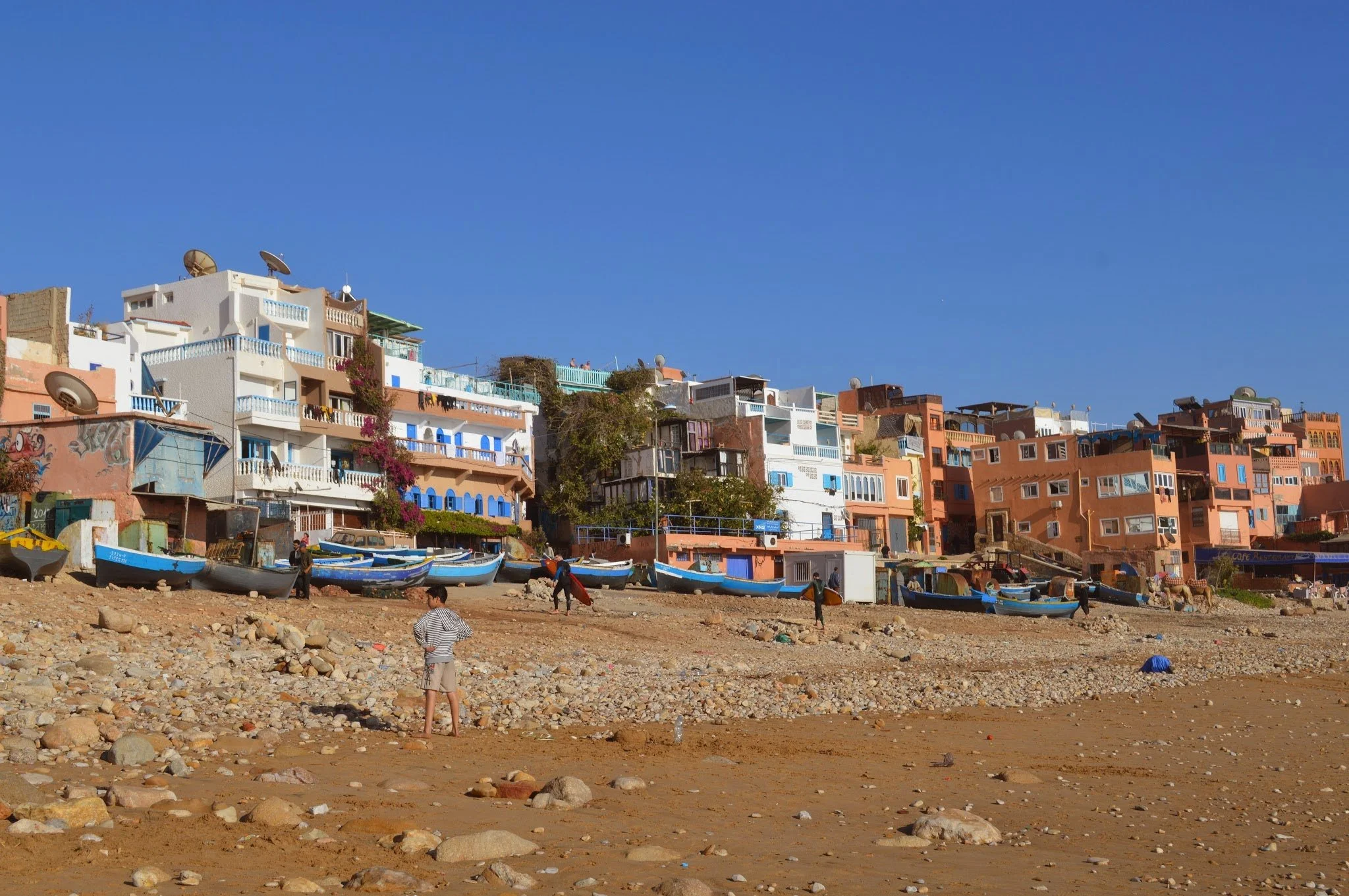 View of colorful houses built on a hillside near a rocky beach, with boats on the shore and a clear blue sky.