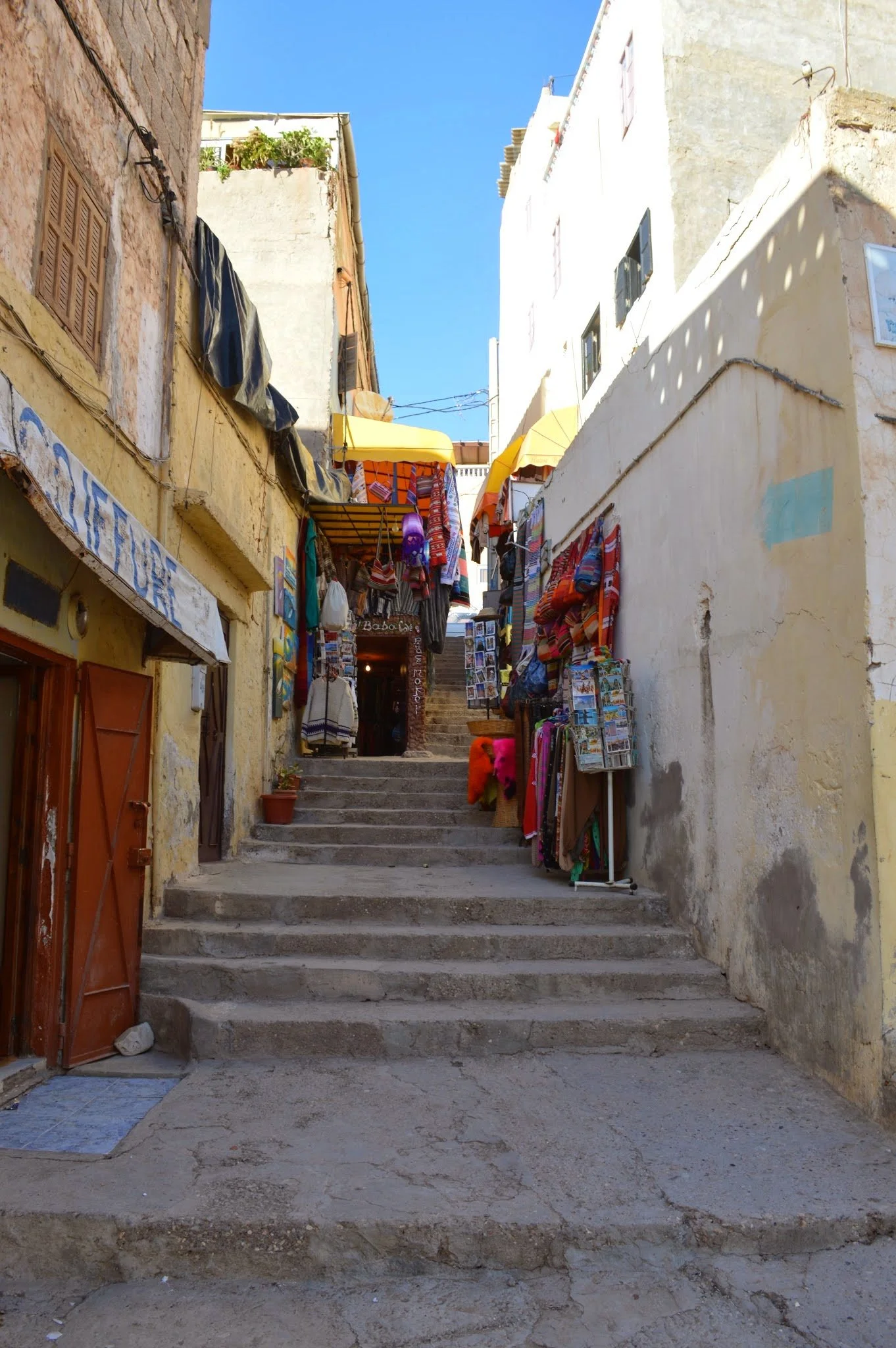 A narrow alleyway with stairs leading upward, lined with shops selling textiles, souvenirs, and postcards. The buildings are old with yellow and white walls, some with visible wear and cracks. Umbrellas and awnings hang overhead, providing shade.