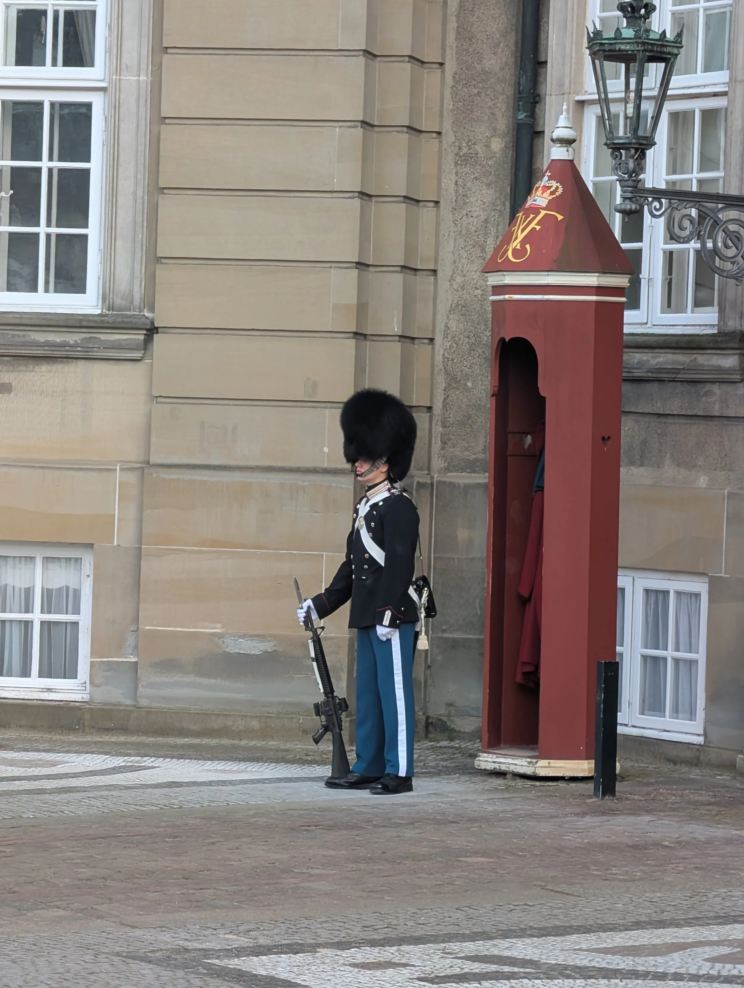 A guard in a formal uniform with a tall bearskin hat standing next to a red British postal box on a cobblestone street in front of a beige building with white window frames.