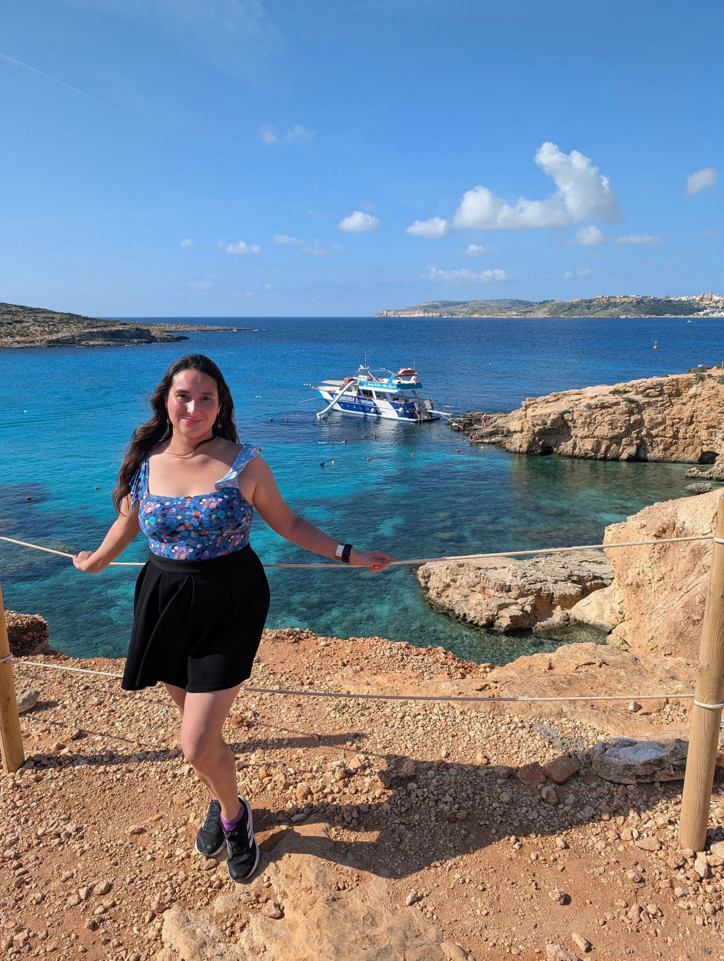 A young woman with long dark hair standing on rocky shore near the water, wearing a blue patterned top, black skirt, and sneakers, with a boat and blue ocean in the background under a partly cloudy sky.