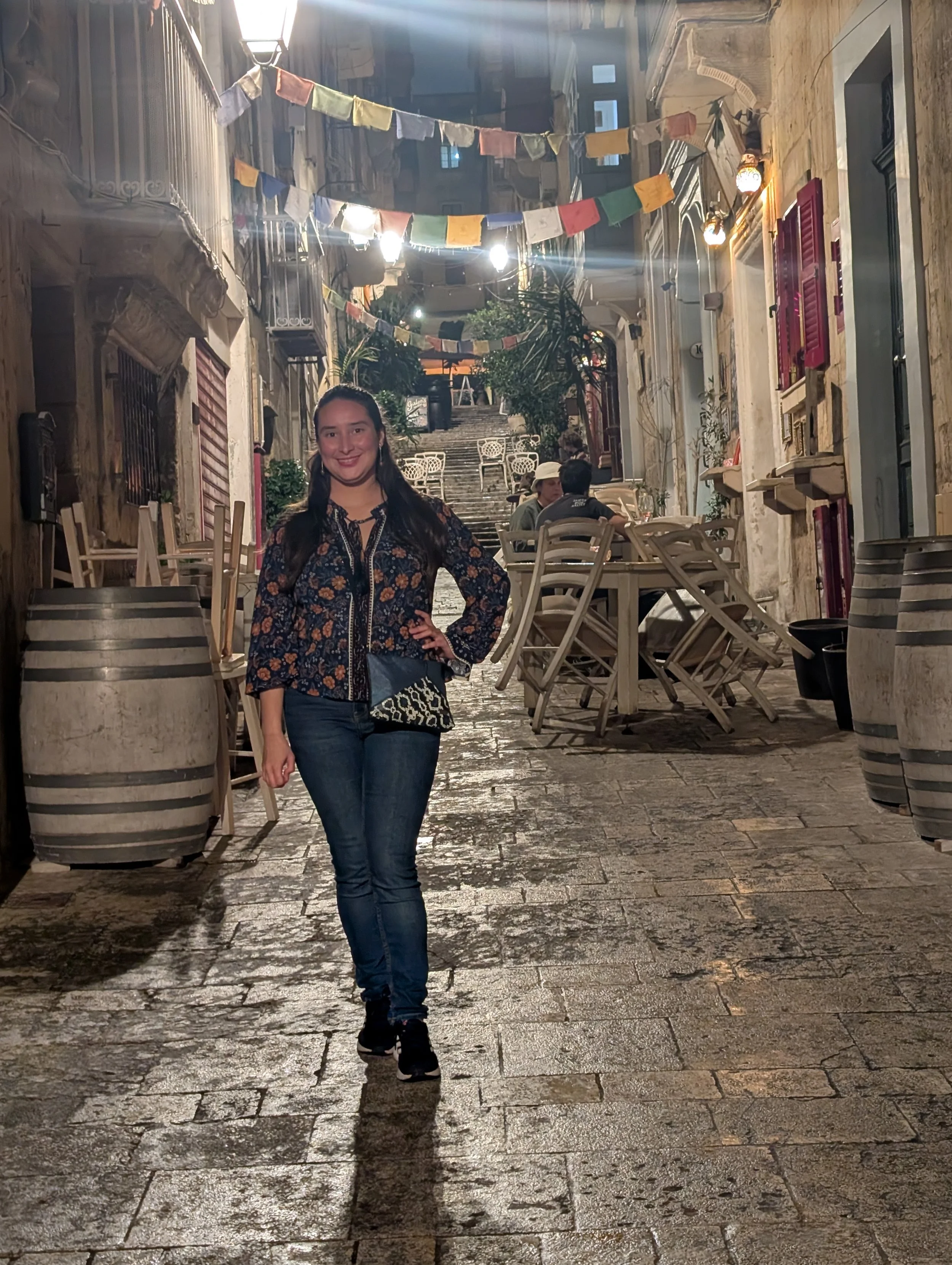 A woman with dark hair wearing a floral blouse and jeans, smiling and standing in a European-style outdoor alley at night, decorated with colorful banners, with a staircase and buildings visible in the background.