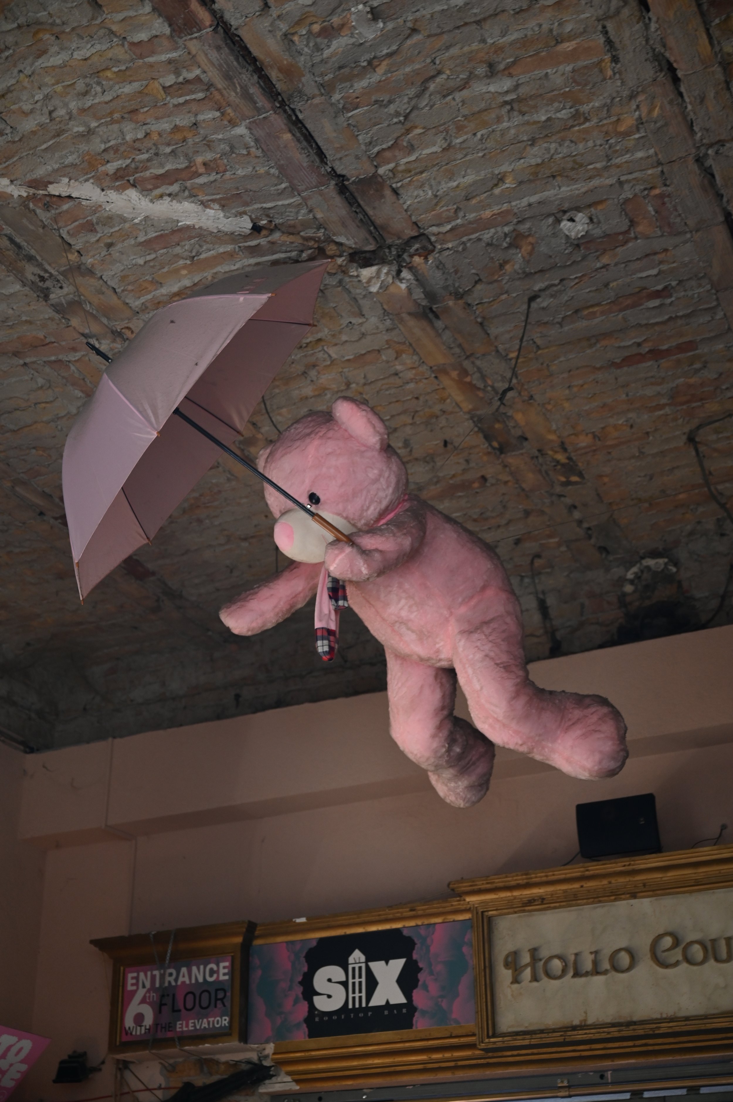 A pink plush teddy bear holding a small light pink umbrella hanging upside down from a brick ceiling. The background features signs indicating the entrance and a store called "Six" and another sign saying "Hello Count".