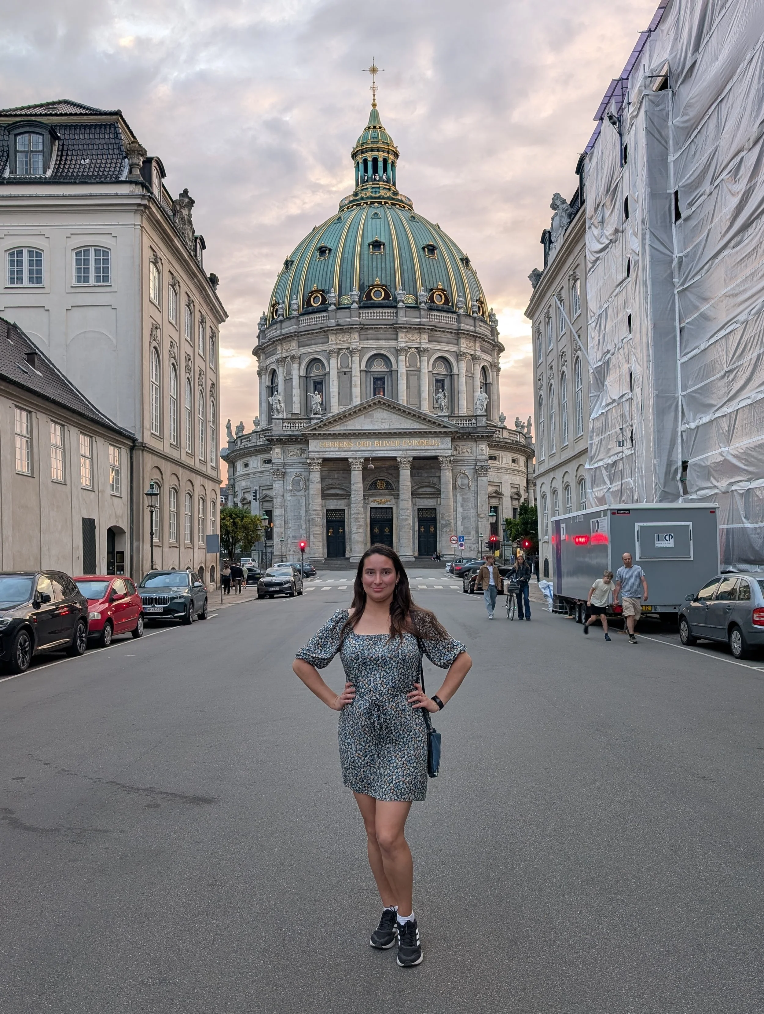 A woman in a patterned dress and sneakers standing on a city street in front of a historic building with a large dome, flanked by two buildings, some parked cars, and people walking.