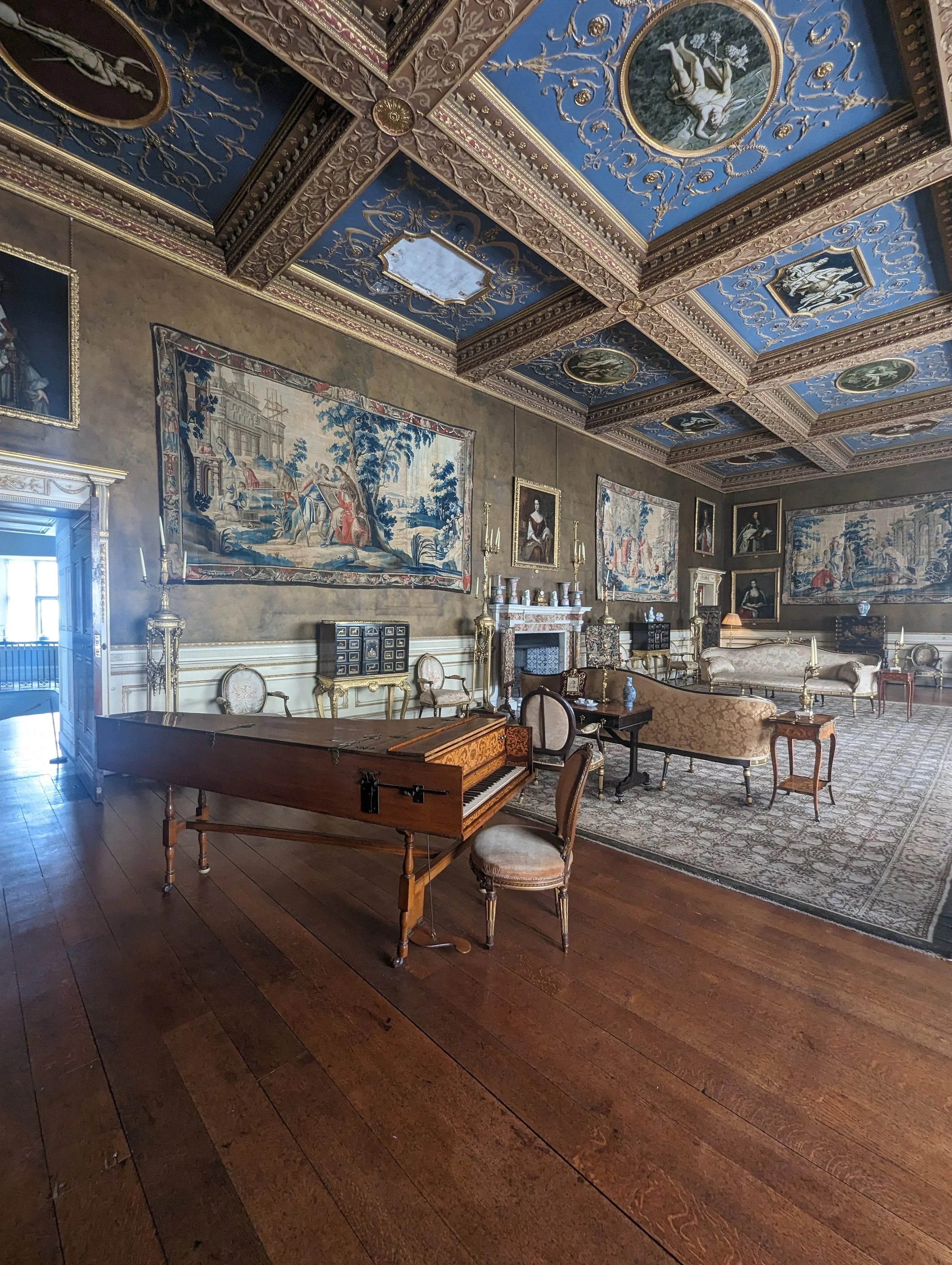 A luxurious room with ornately decorated ceiling featuring paintings and gold accents. The walls are adorned with tapestries and portraits, and the room contains elegant antique furniture including a grand piano, chairs, and tables.