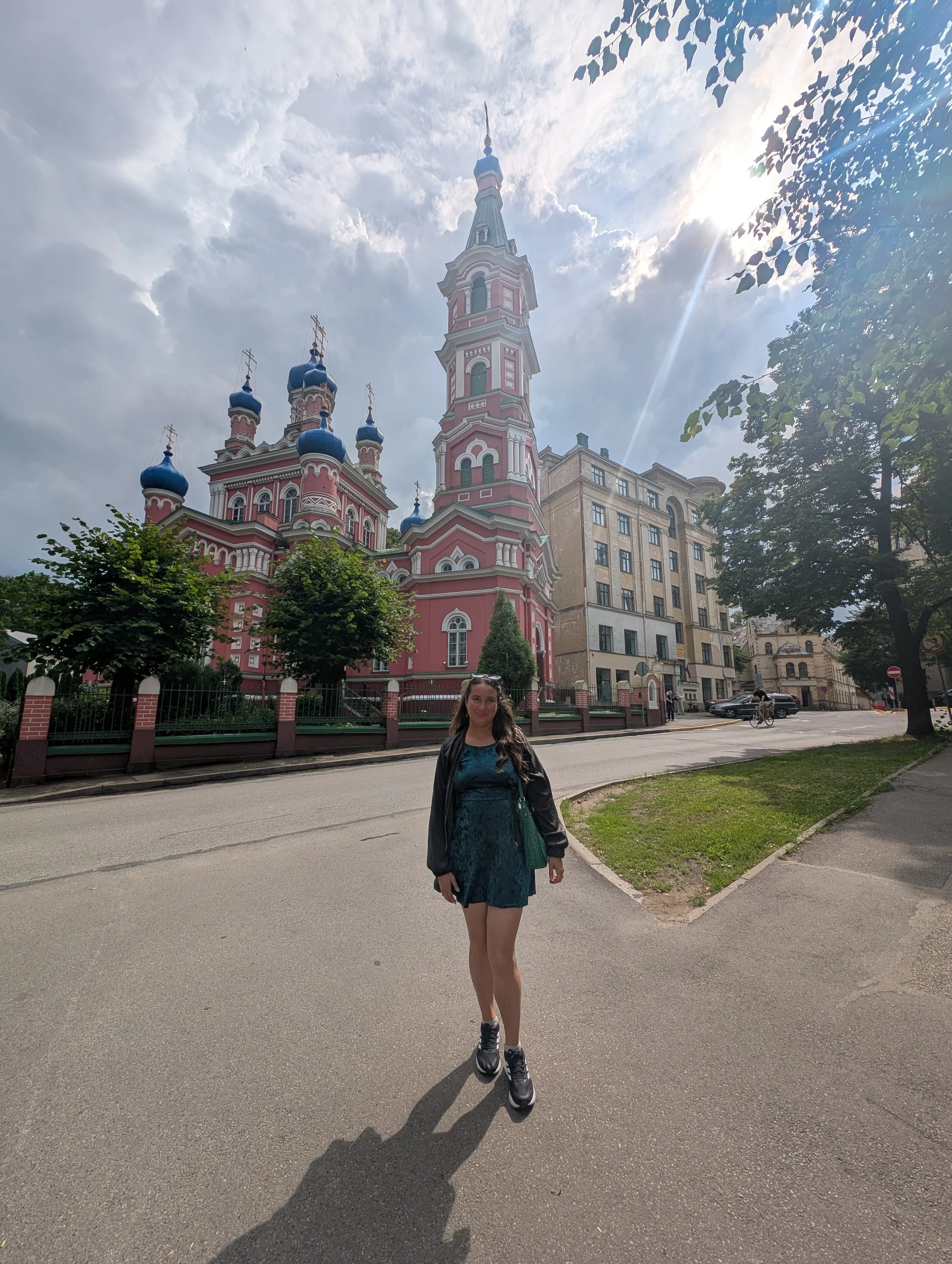 A woman standing on a paved street in front of a pink and white church with blue domes and golden crosses. The sky is partly cloudy with the sun shining through, and there are trees and buildings surrounding the church.