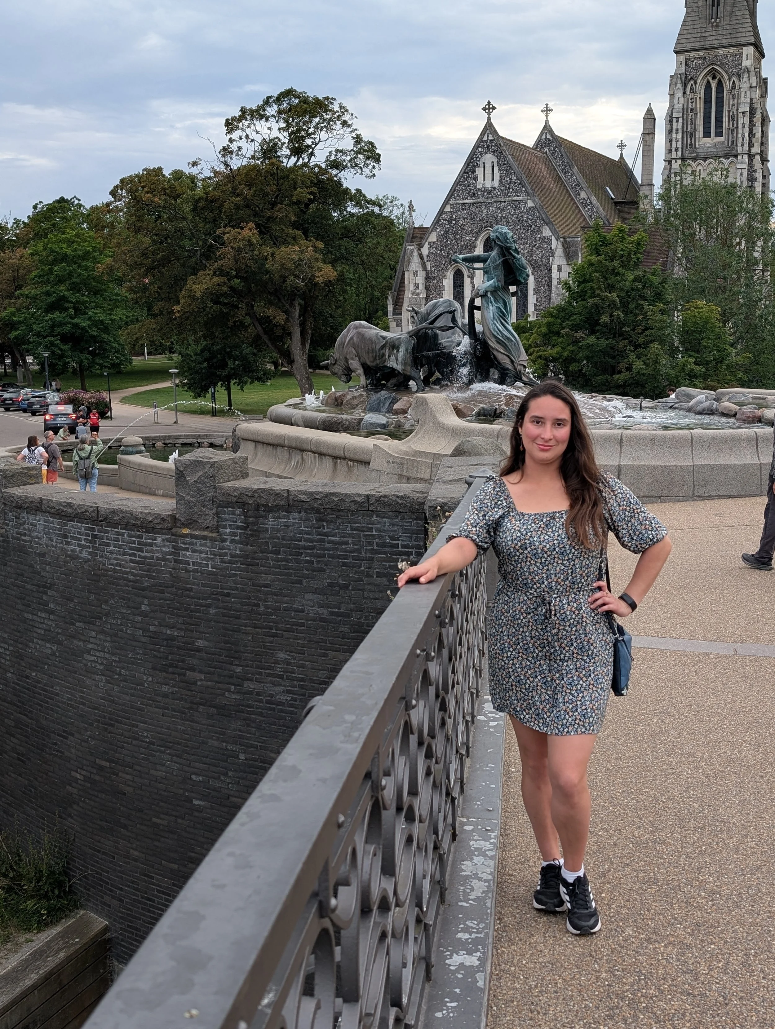 Young woman with long brown hair wearing a floral dress and black sneakers, standing on a bridge with her hand on the railing, smiling in front of a fountain with a statue, trees, a stone church, and a partly cloudy sky in the background.
