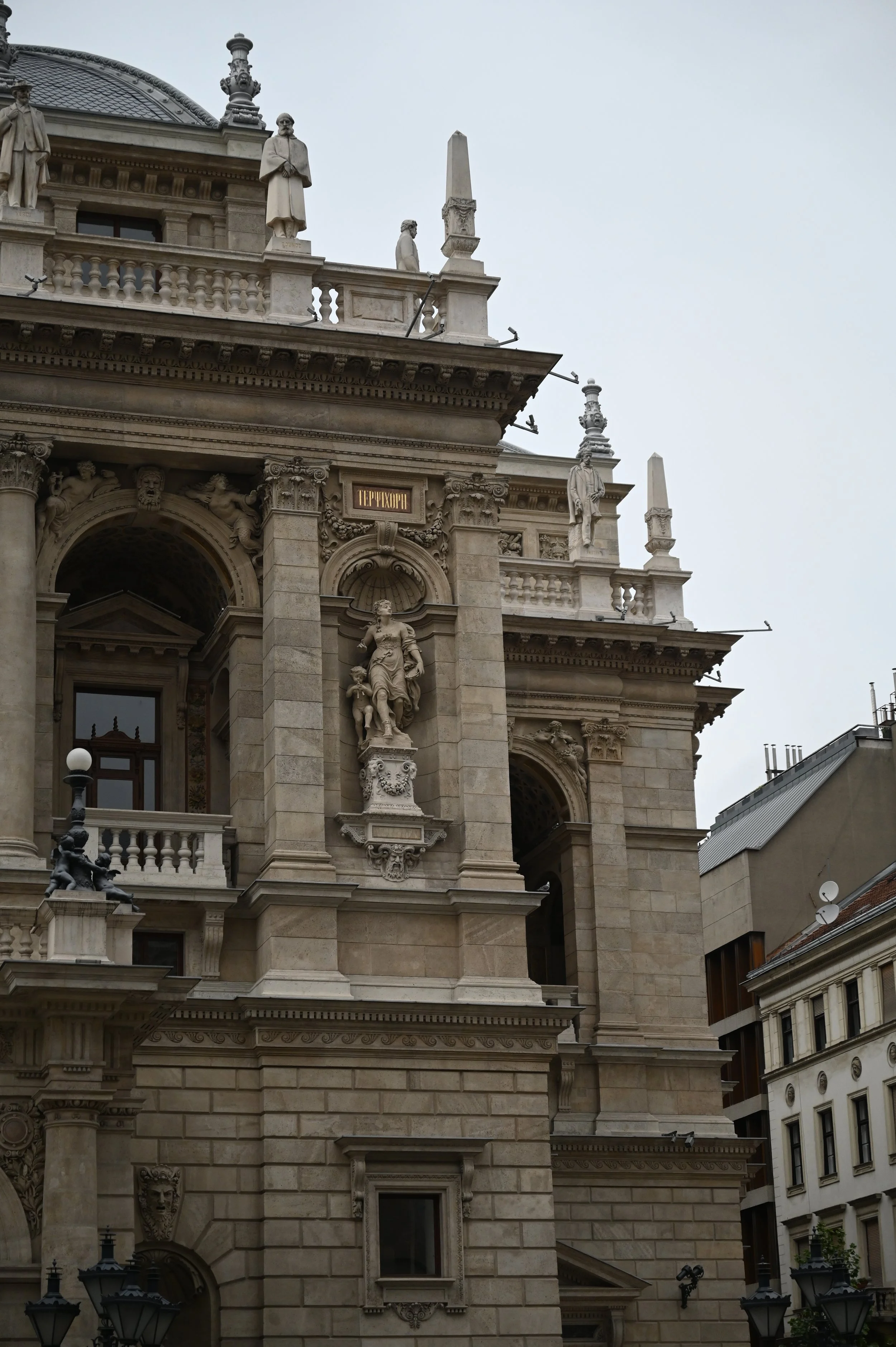 Close-up view of a historic building's ornate facade with sculptures, columns, and decorative elements, including statues on the upper balconies and intricate stonework.