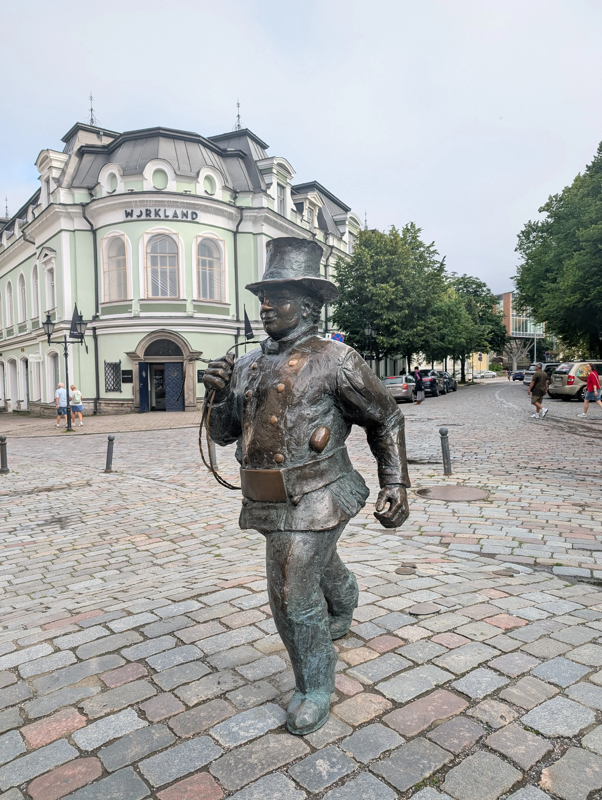 Bronze statue of a man in historical attire, wearing a top hat, holding a pocket watch, and walking on a cobblestone street in front of a historic light green building with the word 'WURKLAND' on it.