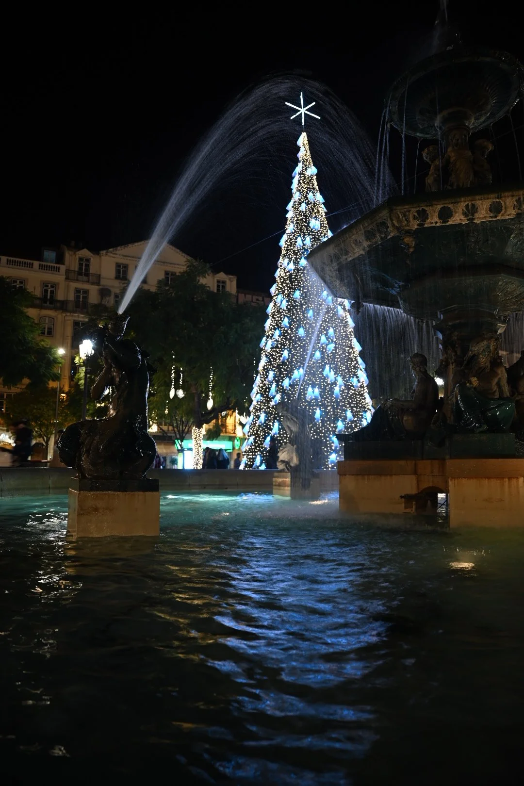 Night scene of a decorated Christmas tree illuminated with blue and white lights, surrounded by ornate fountain statues, with water spraying from the fountain and the tree in a city square.