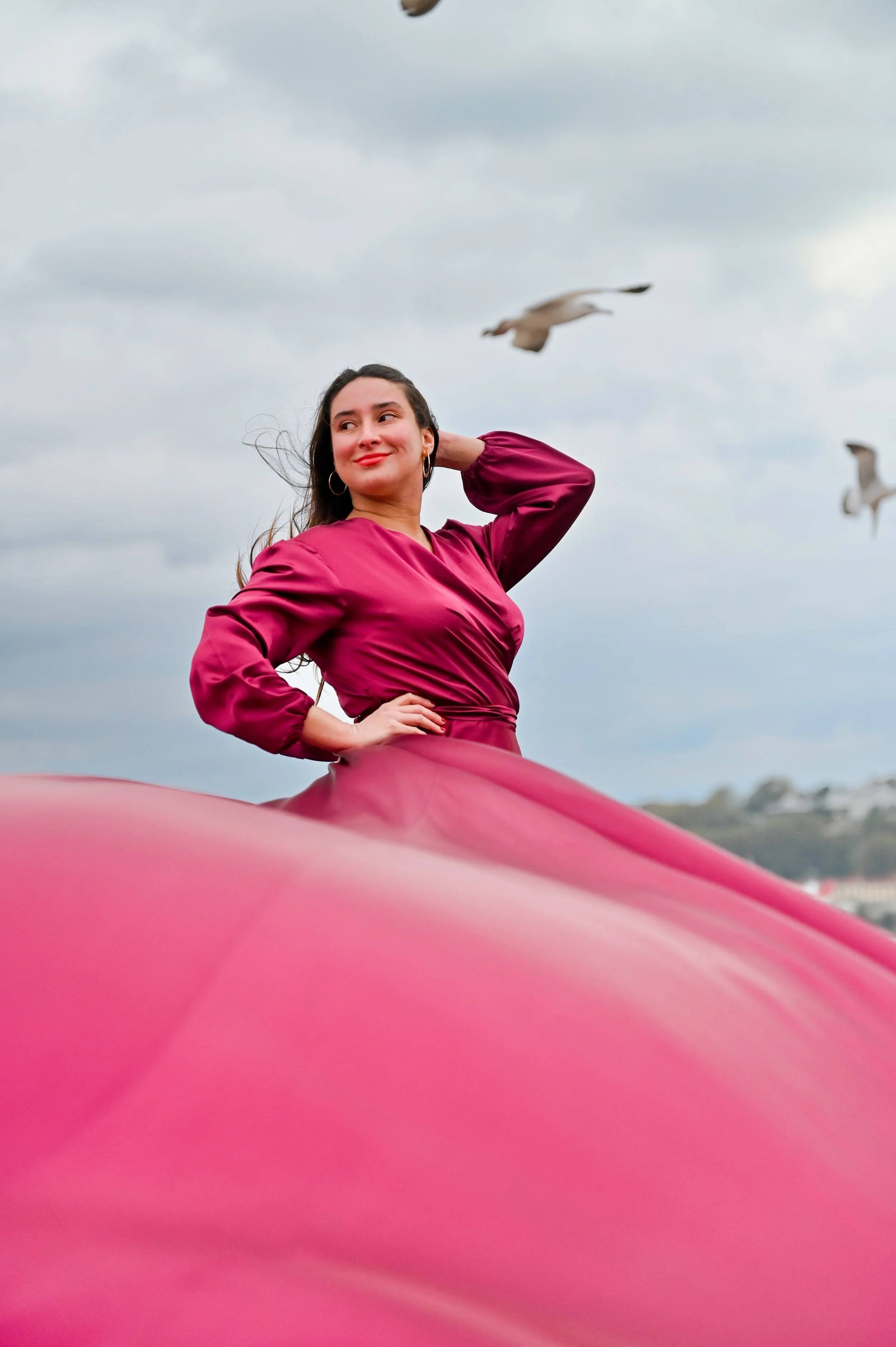 A woman with long dark hair, wearing a purple or magenta dress, smiling with her hand on her head, standing outdoors with a cloudy sky and seagulls flying in the background.