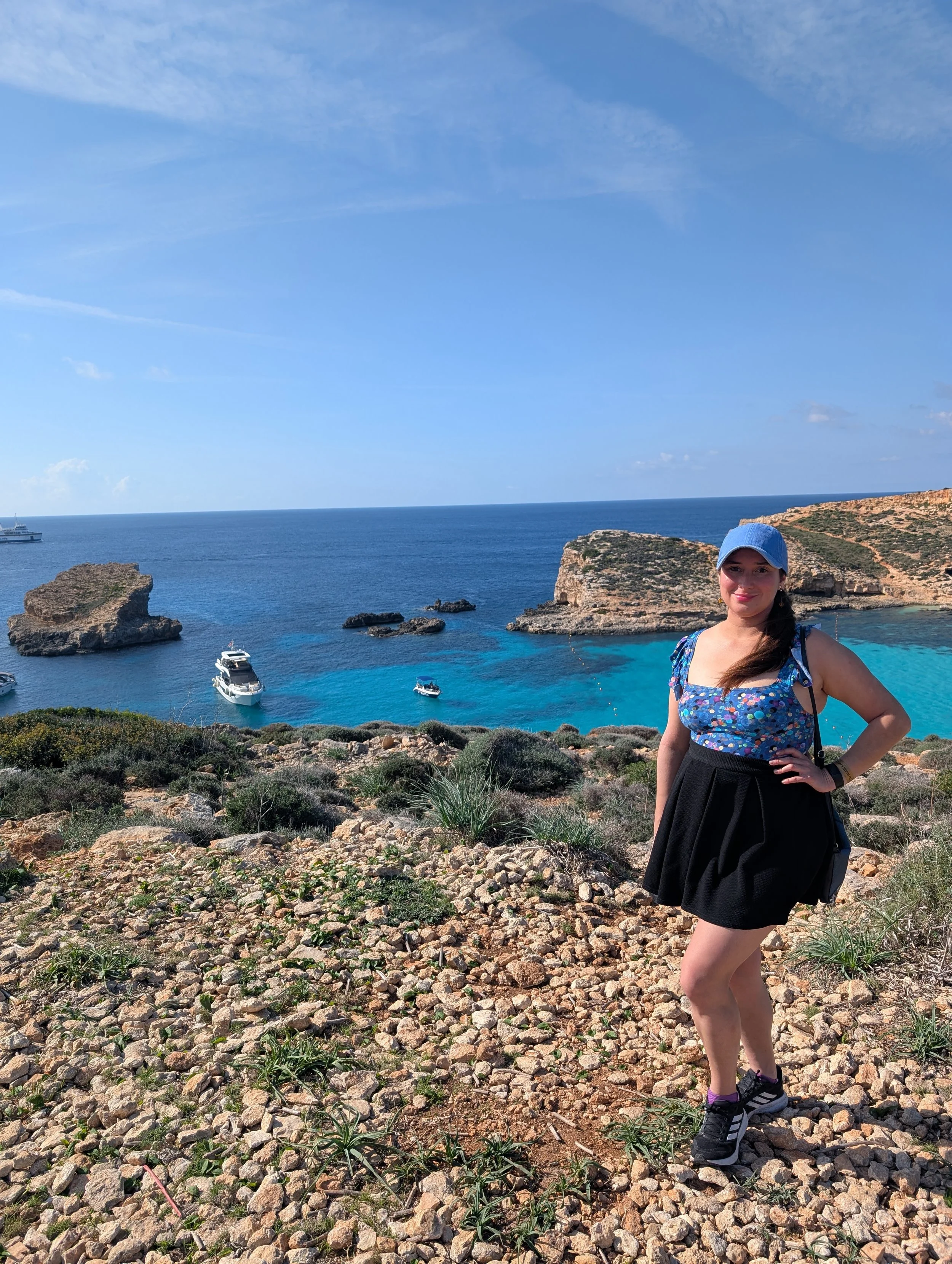 A woman with long dark hair, wearing a blue sun hat, a floral top, a black skirt, purple socks, and black sneakers, standing on a rocky, green hillside overlooking the ocean with boats and rocky cliffs in the background on a sunny day.
