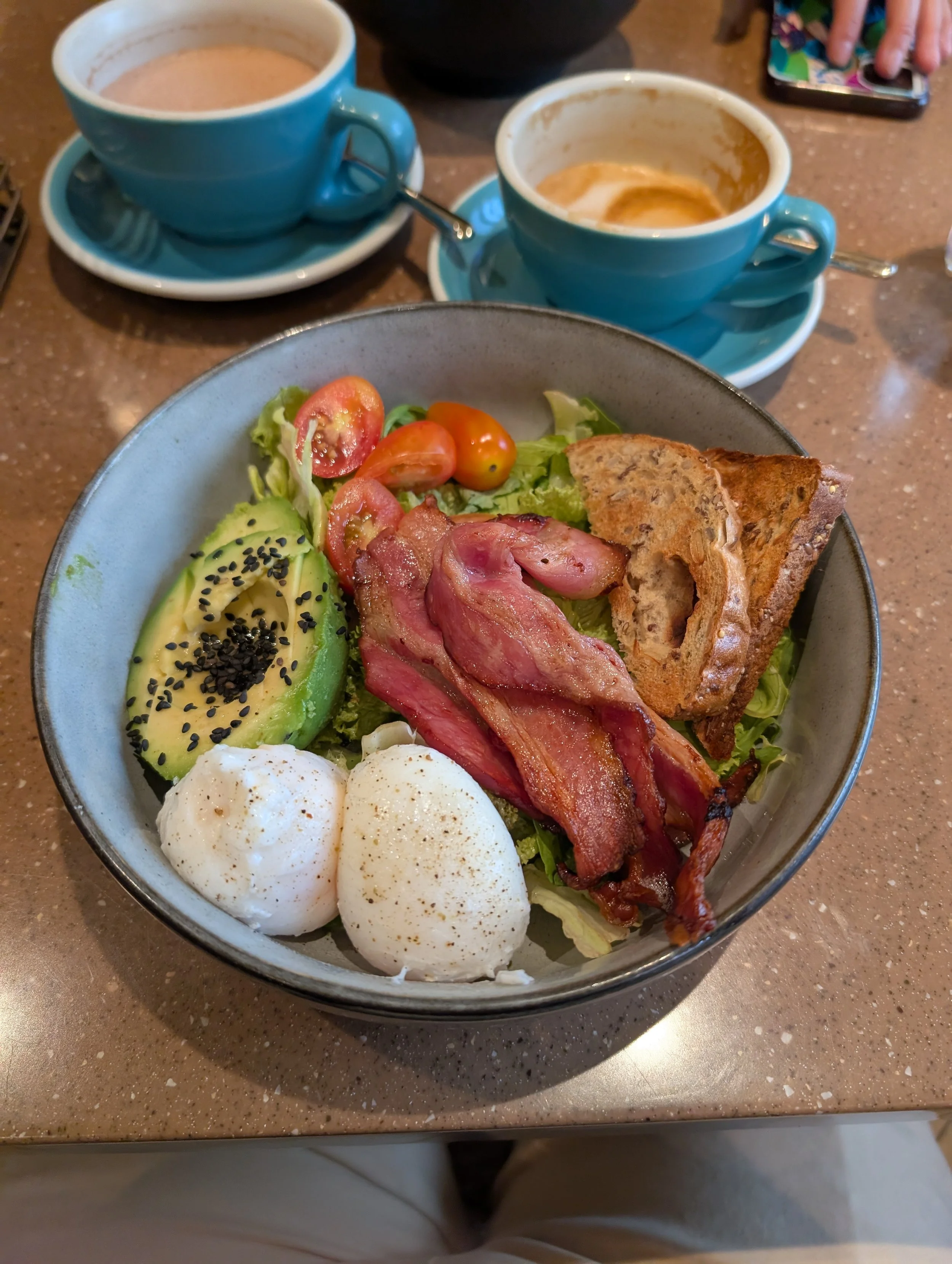 A breakfast plate with poached eggs, sliced avocado sprinkled with black sesame seeds, cherry tomatoes, crispy bacon, and toasted bread. In the background, two cups of coffee and a hand using a smartphone are visible.