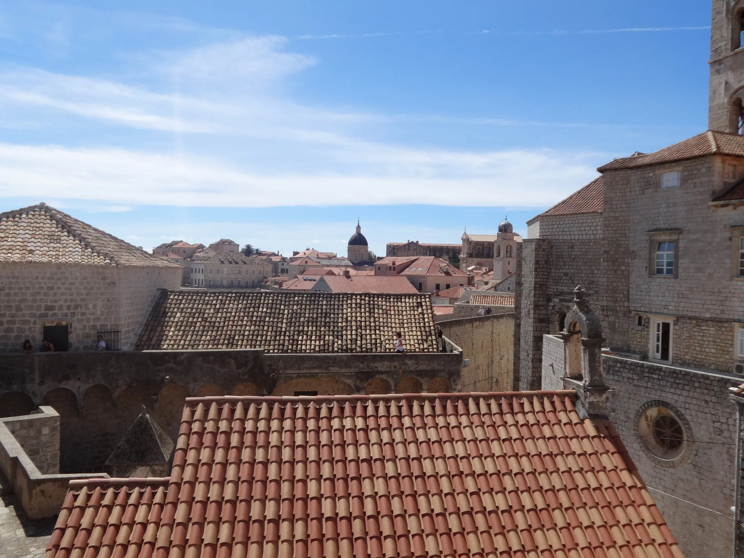 View of old European city with terracotta rooftops, stone buildings, and a church steeple against a blue sky with wispy clouds.