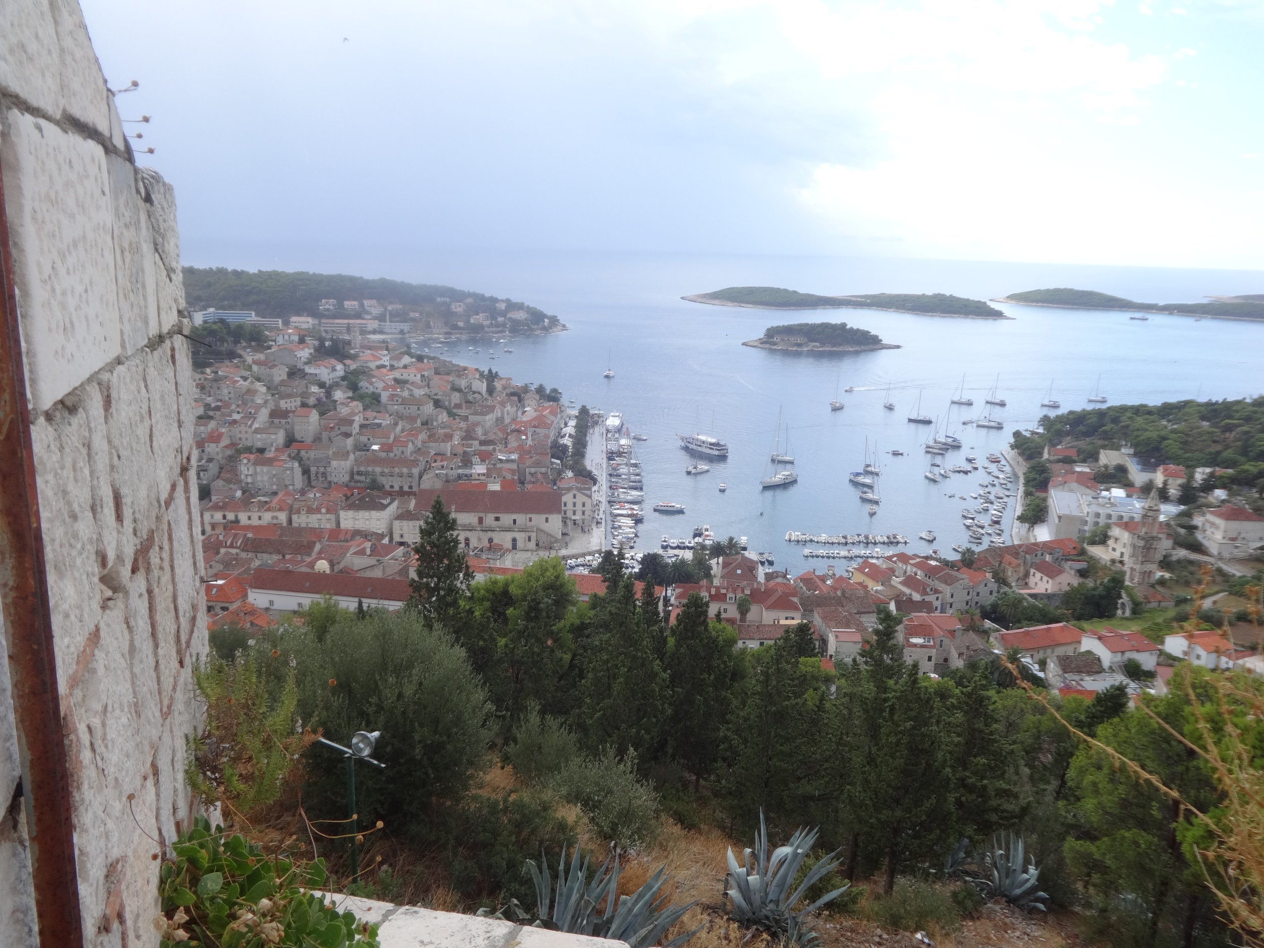 View from a high vantage point overlooking a coastal town with red rooftops, a marina filled with sailboats, and several small islands in the background under a cloudy sky.