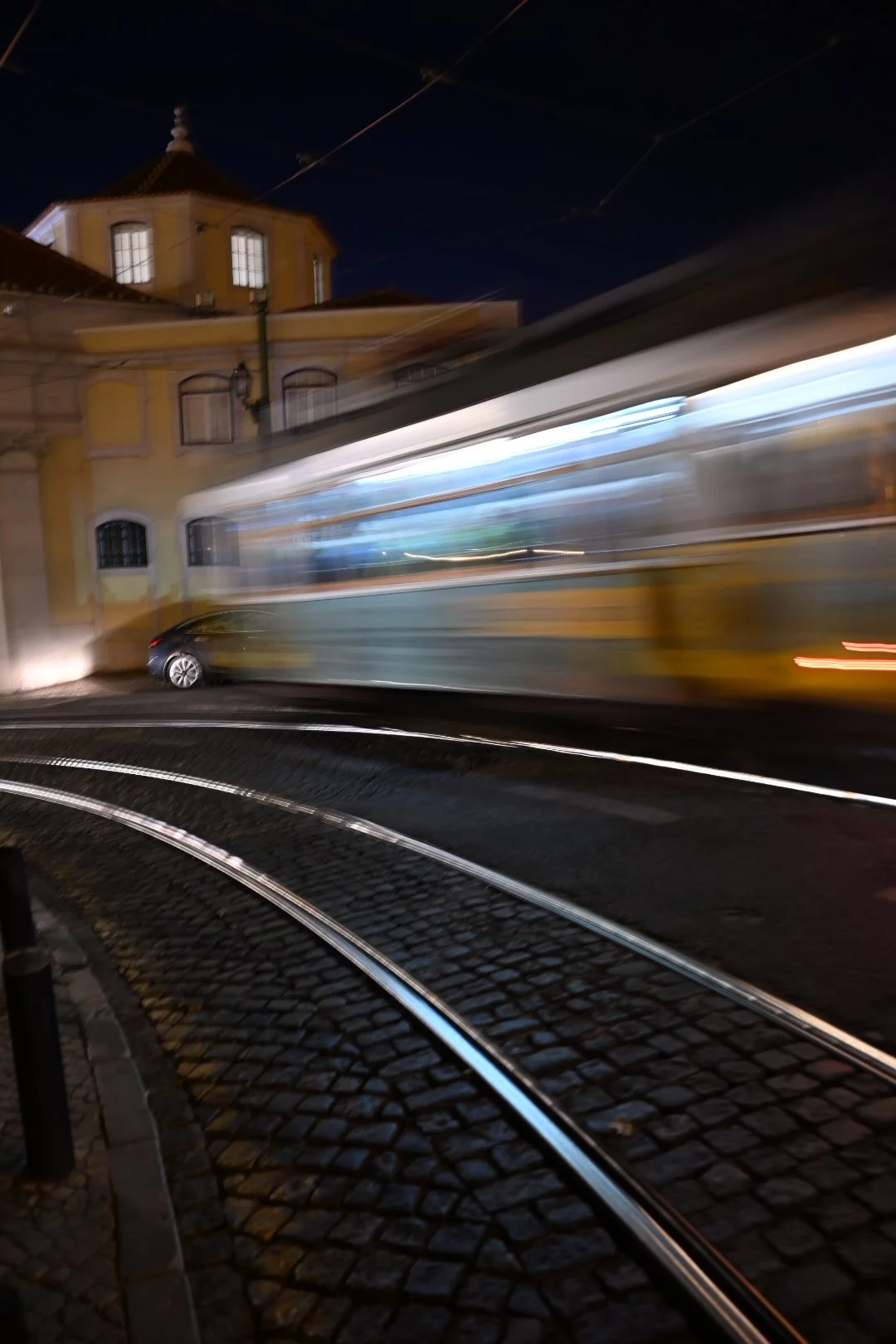 Motion-blurred view of a tram moving past a yellow historic building at night, with tram tracks on cobblestone pavement in the foreground.