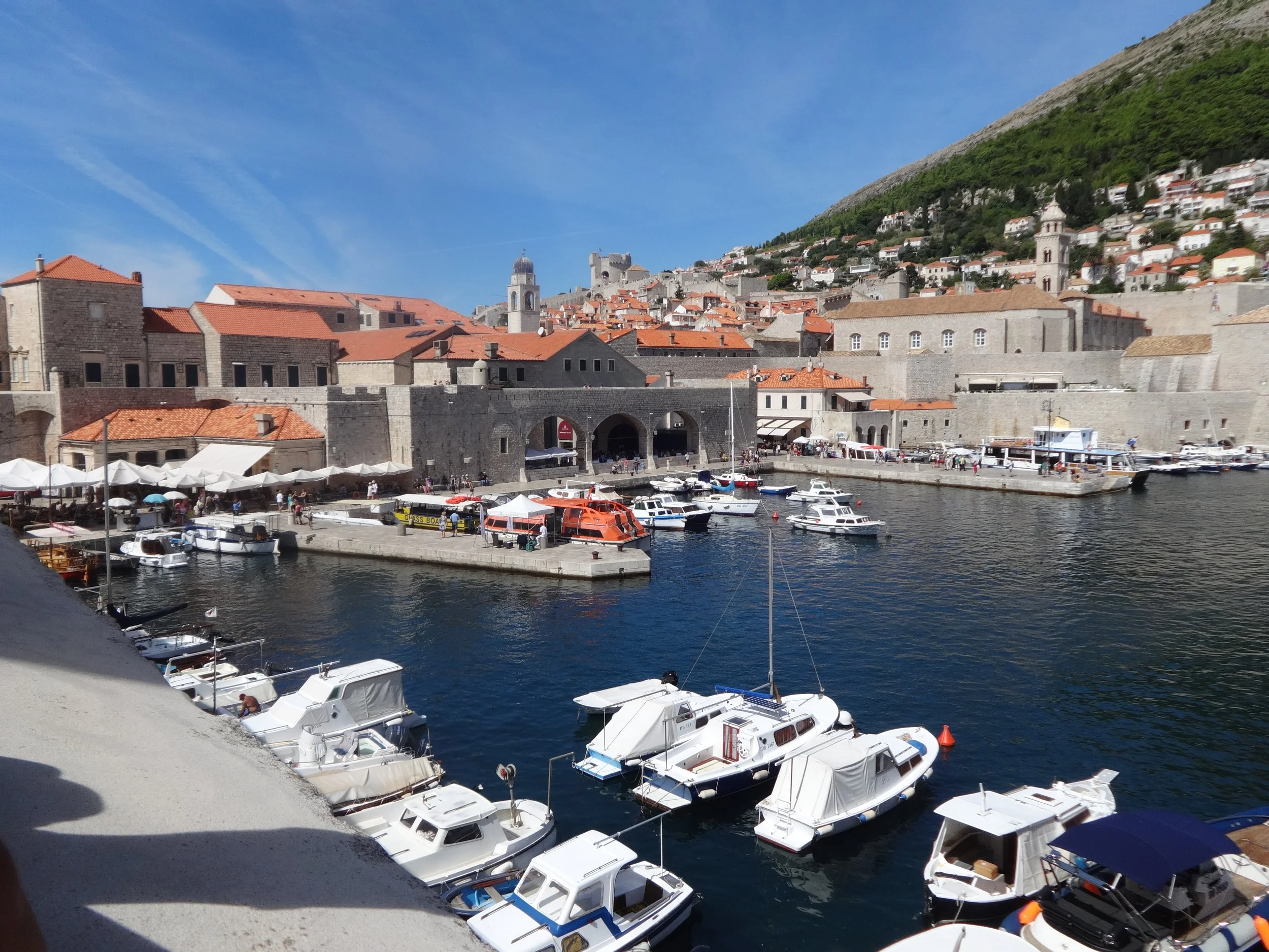 A harbor with numerous small white boats docked along the waterfront, surrounded by stone buildings with orange tiled roofs, a hillside village in the background, a clear blue sky, and a mountain covered in greenery.
