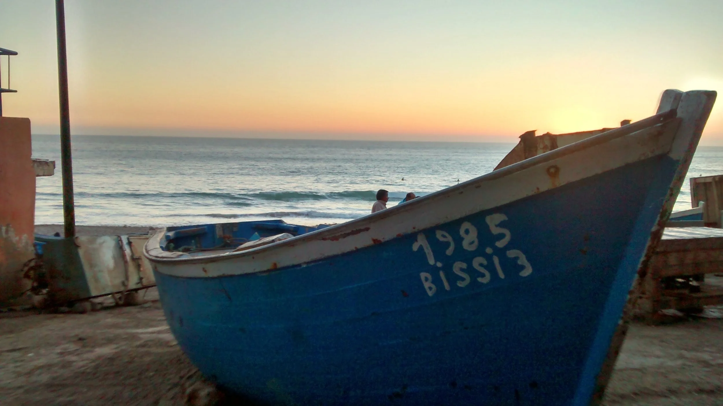 A blue fishing boat with the number 1985 painted on it, resting on a sandy beach at sunset, with the calm ocean and two people in the background.
