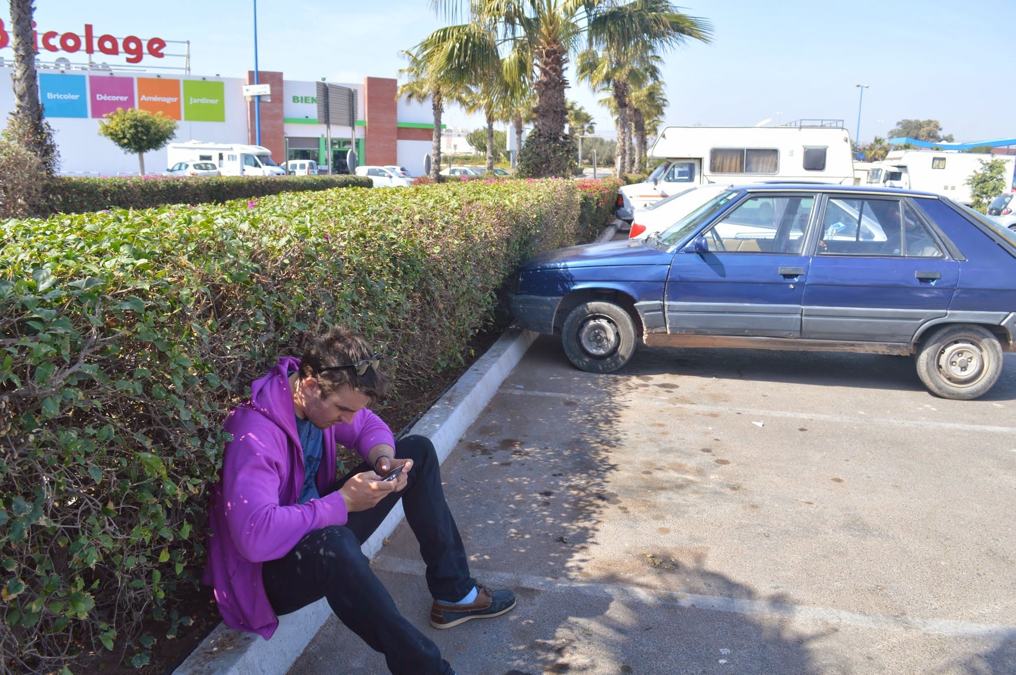 A woman in a purple hoodie and black pants sitting against a bush in a parking lot, looking at her phone, with several parked cars and RVs behind her, and a sign for a home improvement store in the background.
