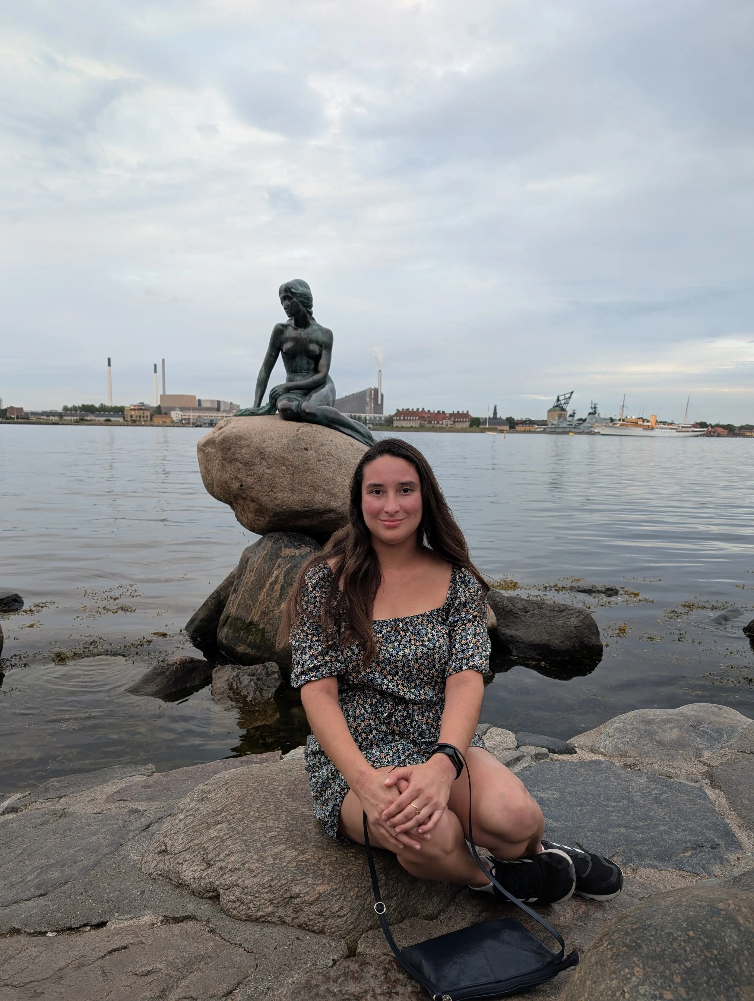 A young woman with long brown hair sitting on rocks by a body of water, with a statue of a reclining mermaid on a large rock behind her. The background includes industrial buildings and ships, with a cloudy sky overhead.