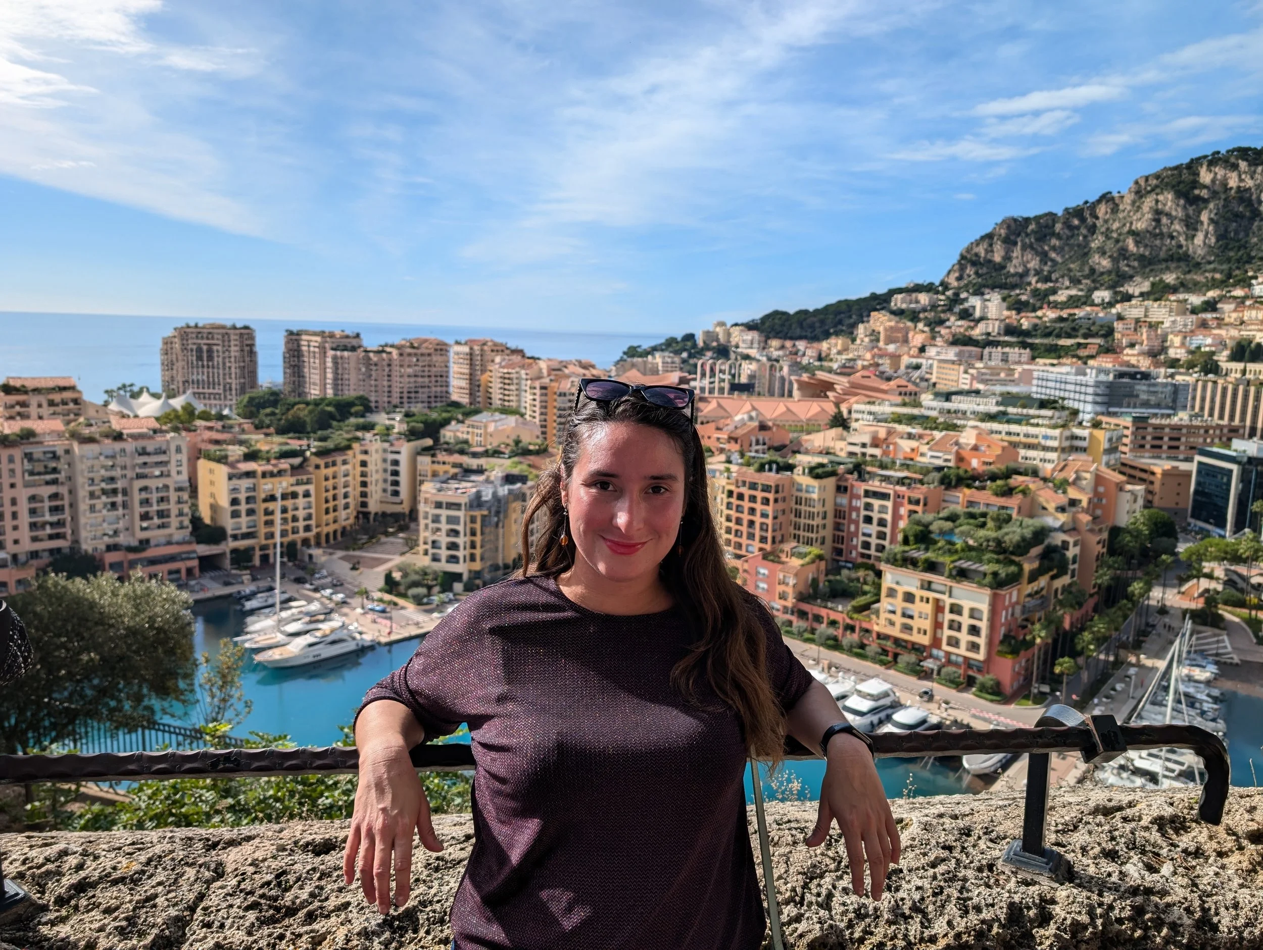 A woman with sunglasses on her head taking a selfie on a balcony with a cityscape and mountains in the background.