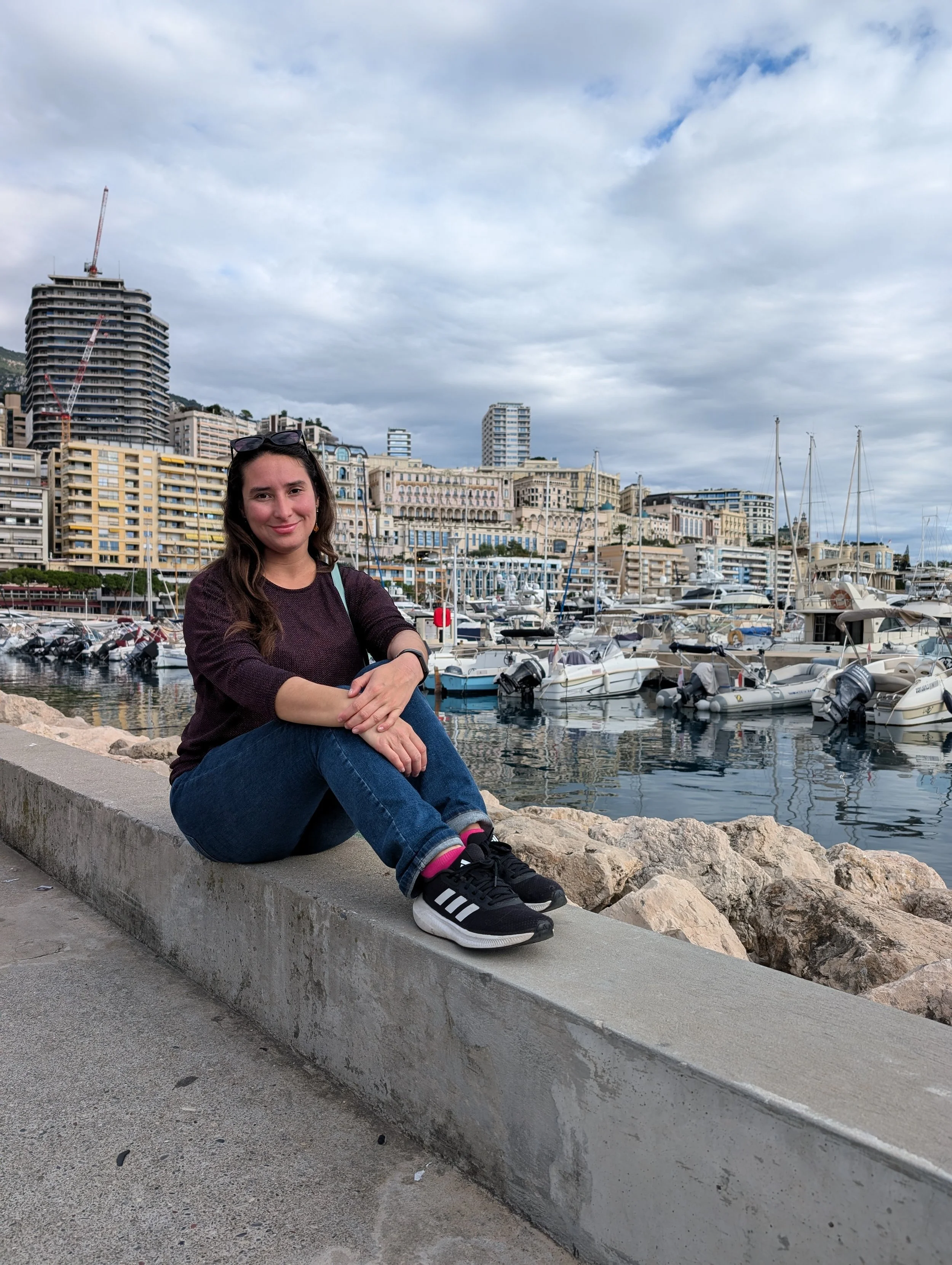 A young woman with long brown hair, wearing a dark sweater, jeans, and sneakers, sitting on a concrete ledge near a marina with sailboats, high-rise buildings, and a cloudy sky in the background.