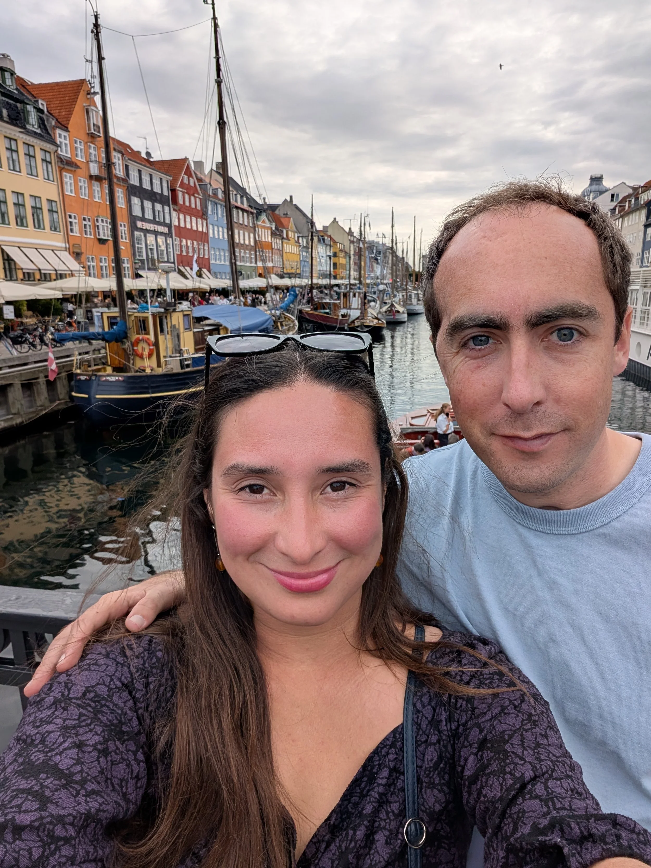 A woman with long brown hair and a man with short light brown hair taking a selfie at a harbor with colorful buildings and boats in the background on a cloudy day.
