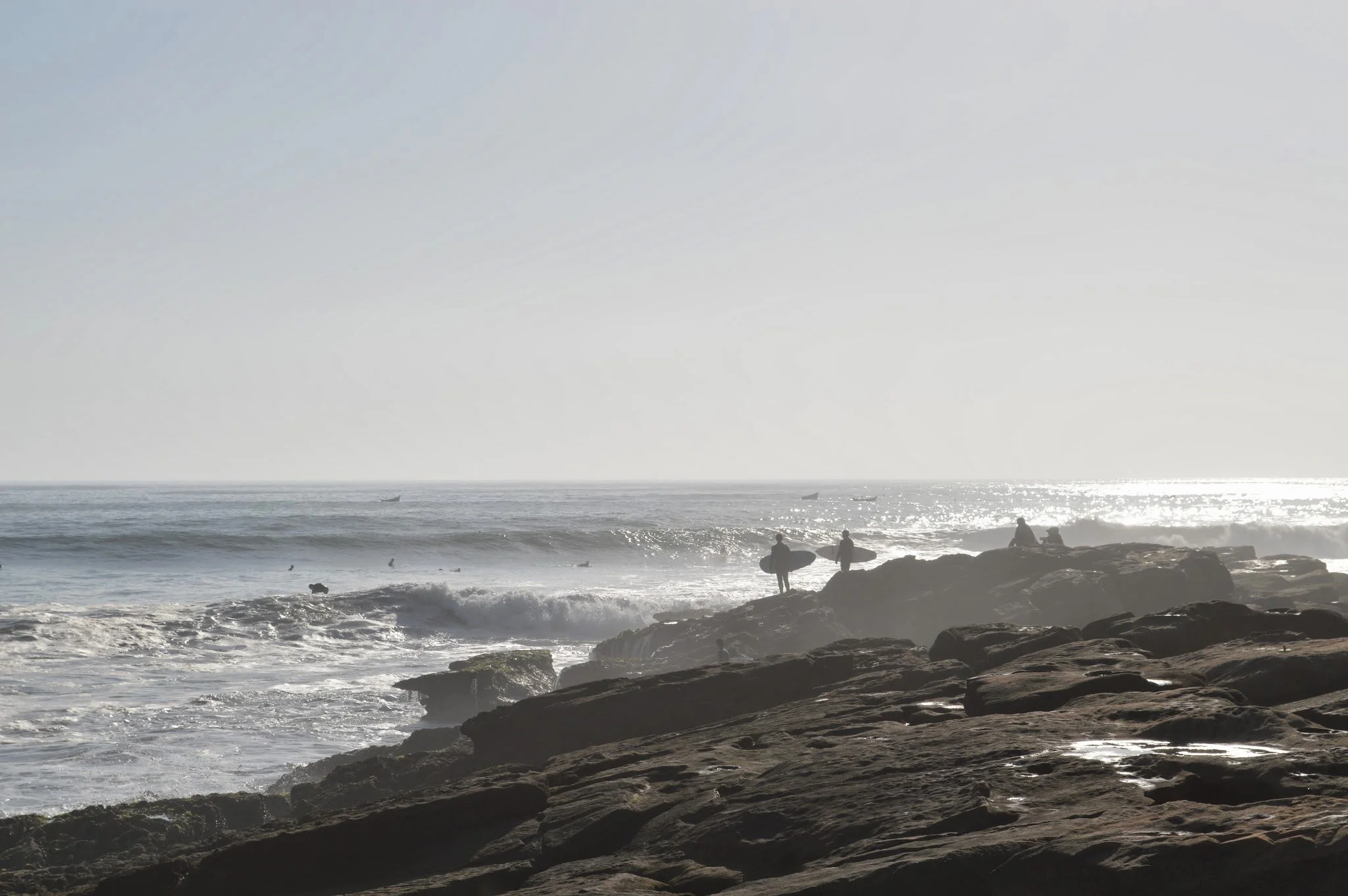 People with surfboards walking on rocky shore near ocean waves at sunset.