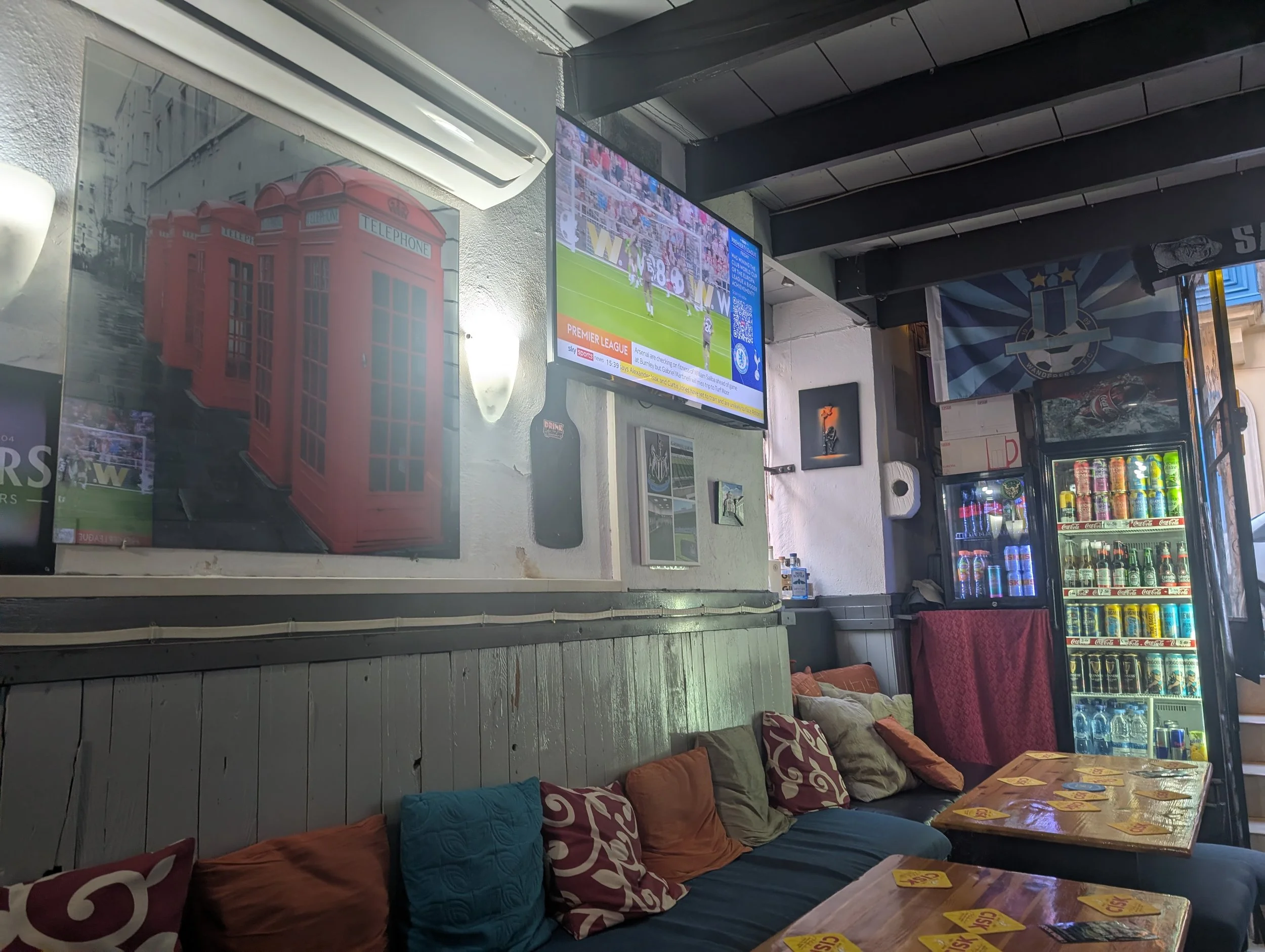 Interior of a room with a television showing a soccer game, a vending machine stocked with drinks, a seating area with colorful cushions, and various posters and decorations on the wall.