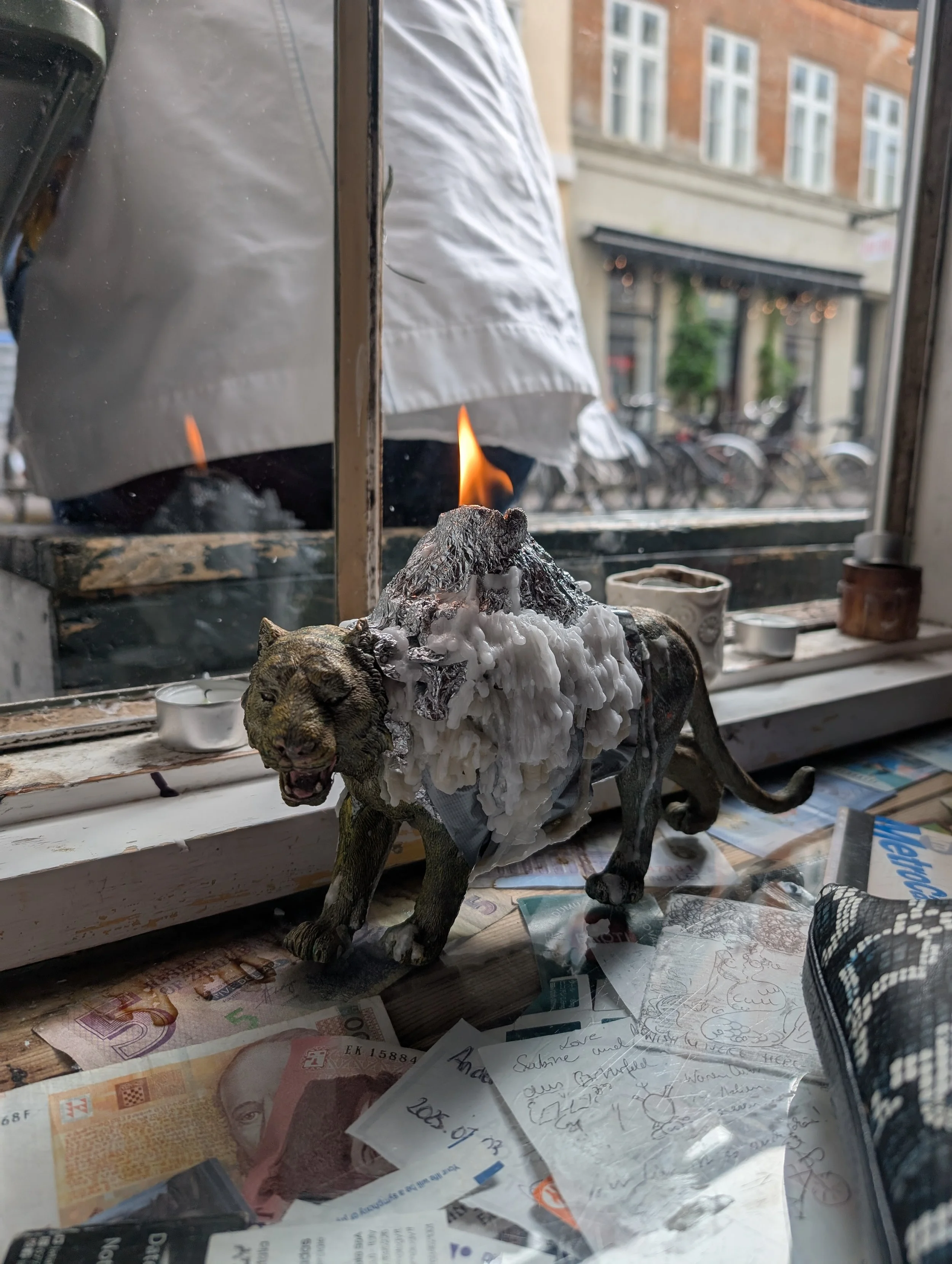 A small model of a wild cat with gray fur is placed on a window sill. The fur on its back is crafted to resemble a mountain with snow, and a small flame is lit on top of the mountain. The window shows an urban street scene outside with bicycles and b