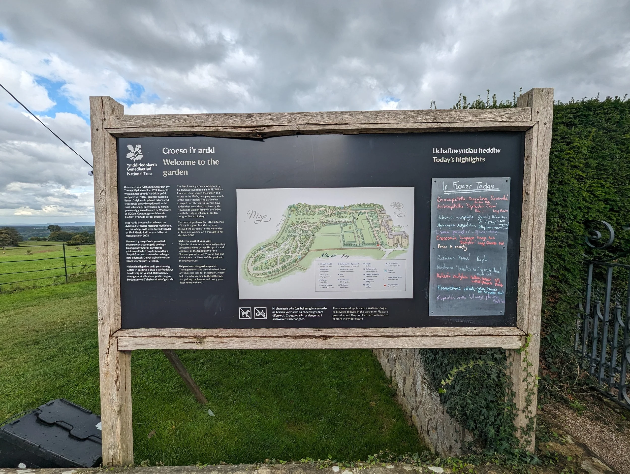 A large outdoor informational sign about a garden, featuring text in English and Welsh, a map of the garden layout, and a list of highlights for the day, set against a scenic rural landscape with cloudy sky and green fields.