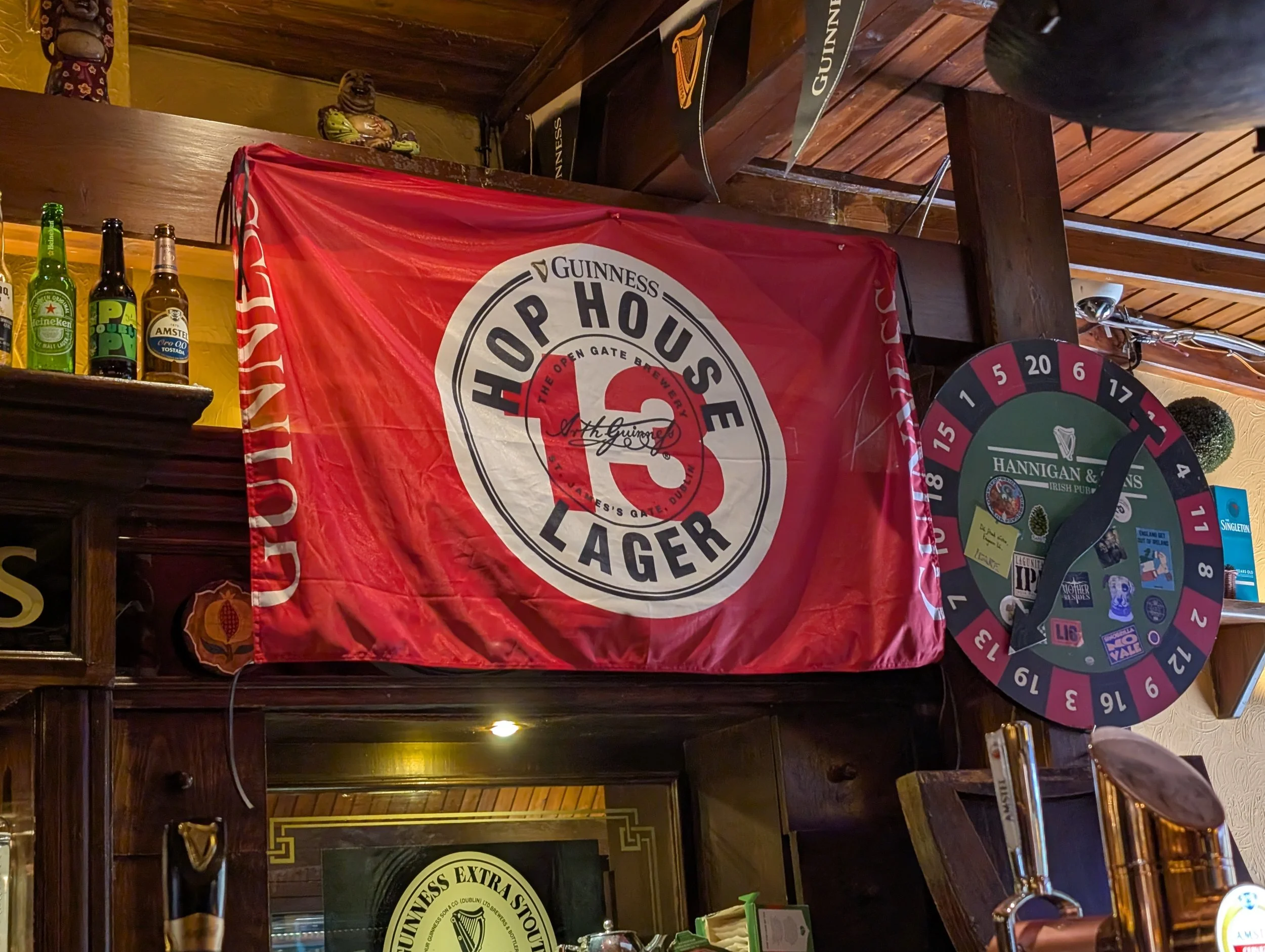Bar area with Guinness flag, various beer bottles, a dartboard, and signage in a pub or restaurant setting.