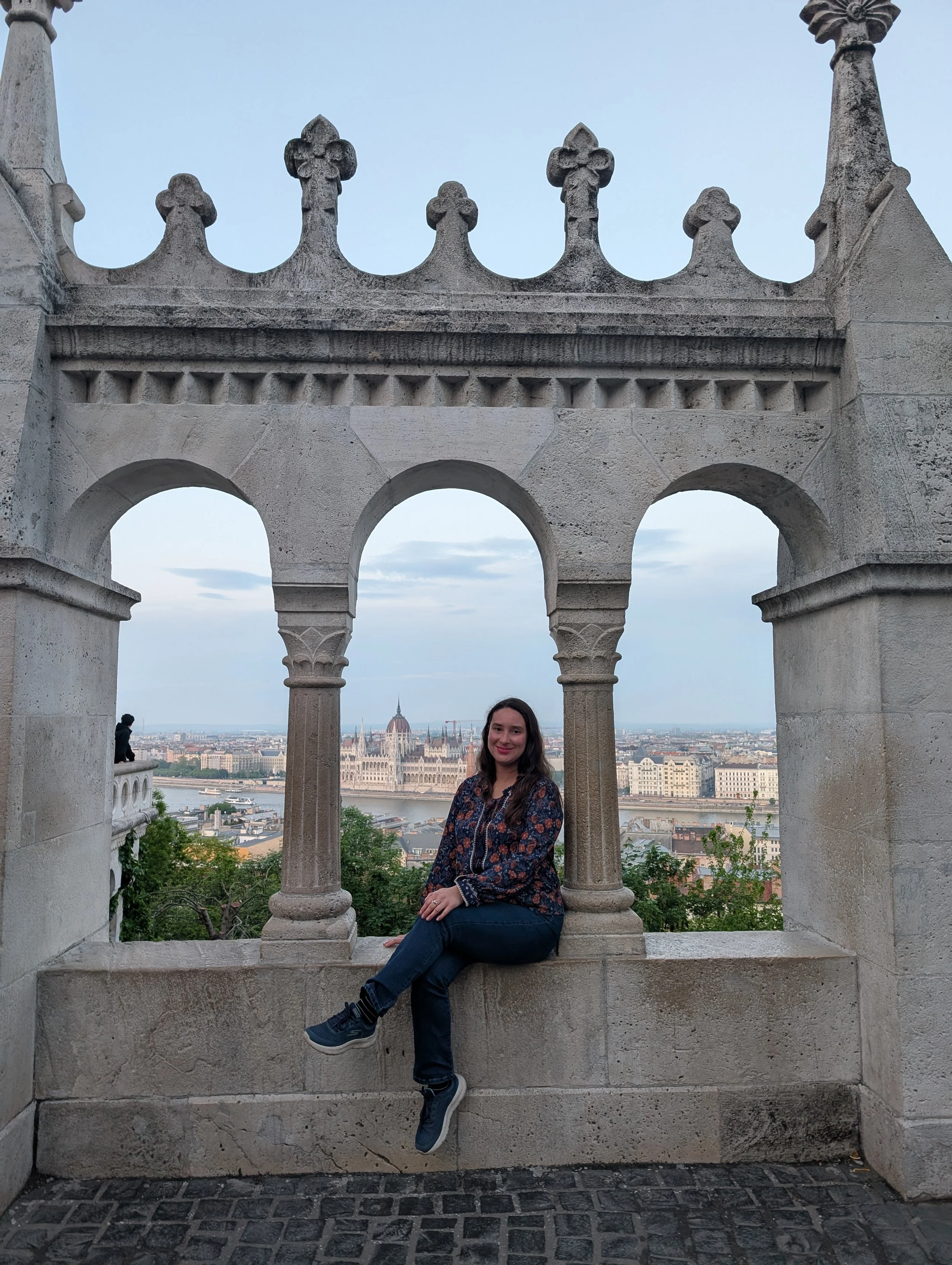 Woman sitting on a stone ledge in front of an ornate stone archway overlooking the city, including the Hungarian Parliament Building across the Danube River in Budapest.