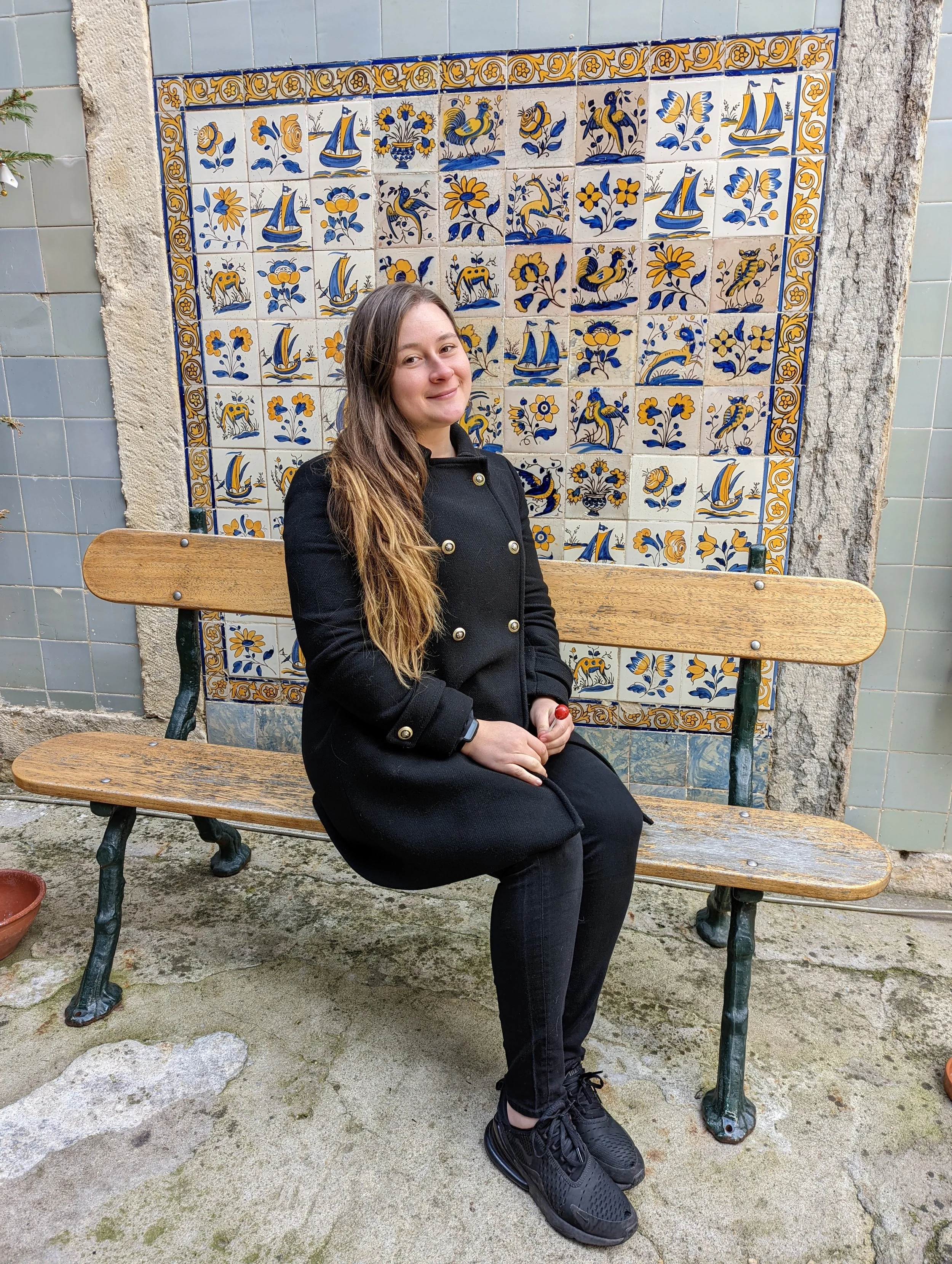 A young woman with long brown hair sitting on a wooden bench in front of a decorative tile wall. She is wearing a black coat and black sneakers, smiling slightly. The background features a tiled wall with blue and yellow designs, and the ground is un