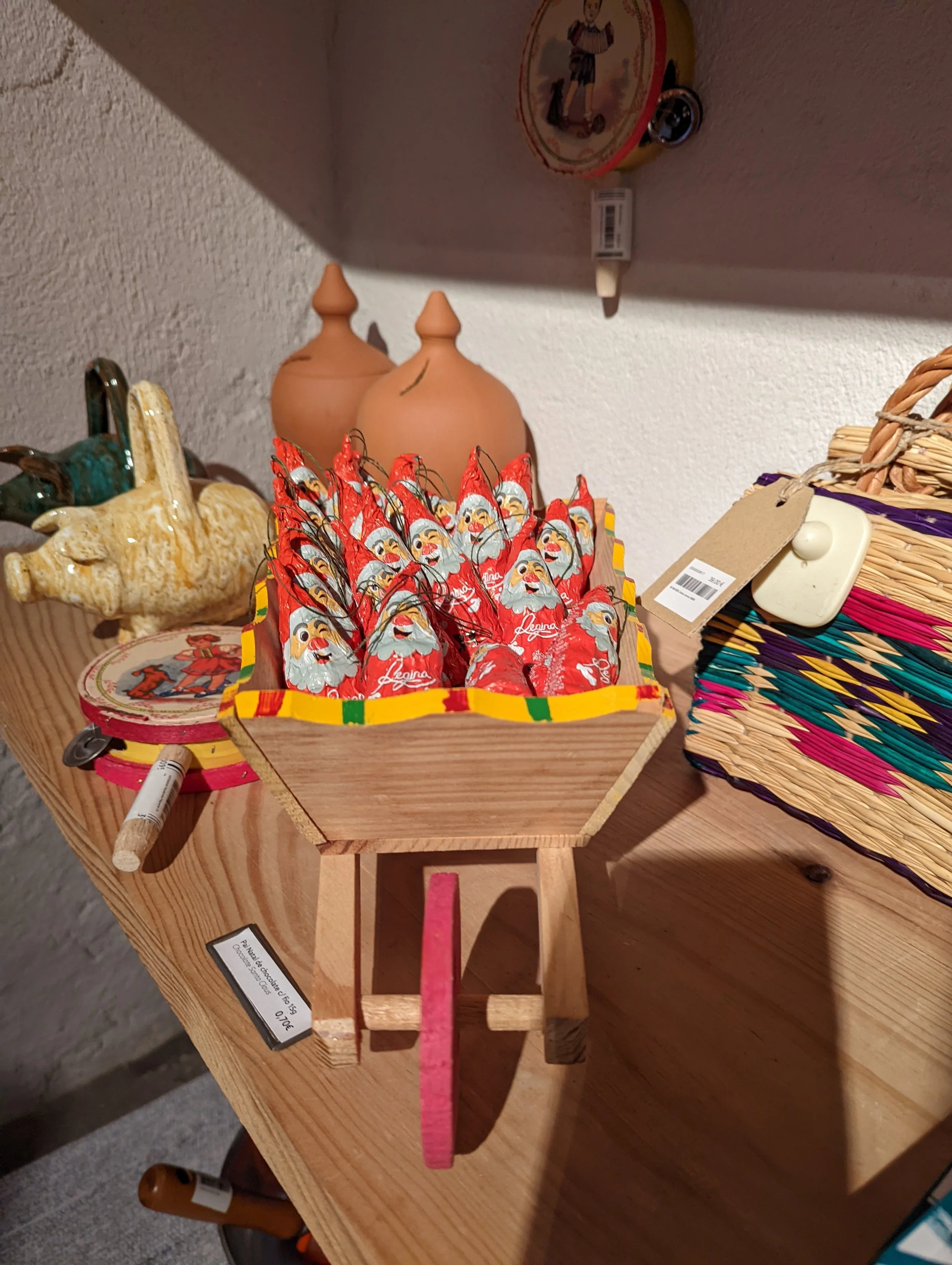 Decorative Santa Claus chocolate candies in red wrappers arranged in a tray, a pair of ceramic jugs behind, a small round coaster, and a woven basket on the right side, all placed on a wooden table.