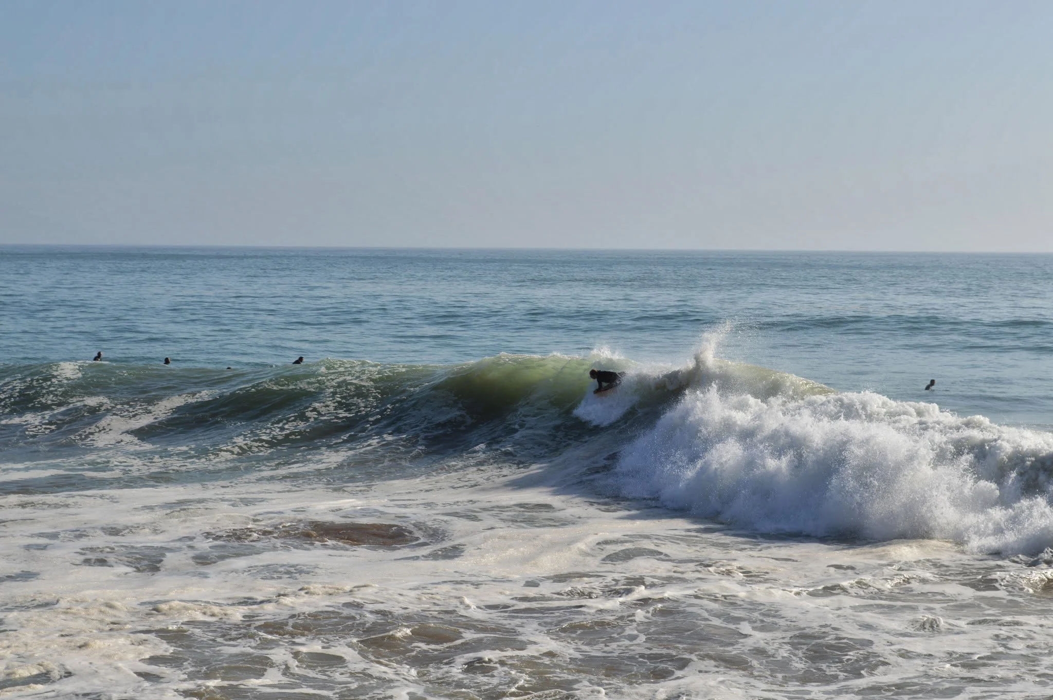 The image shows the ocean with gentle waves and several people surfing in the water under a clear sky.