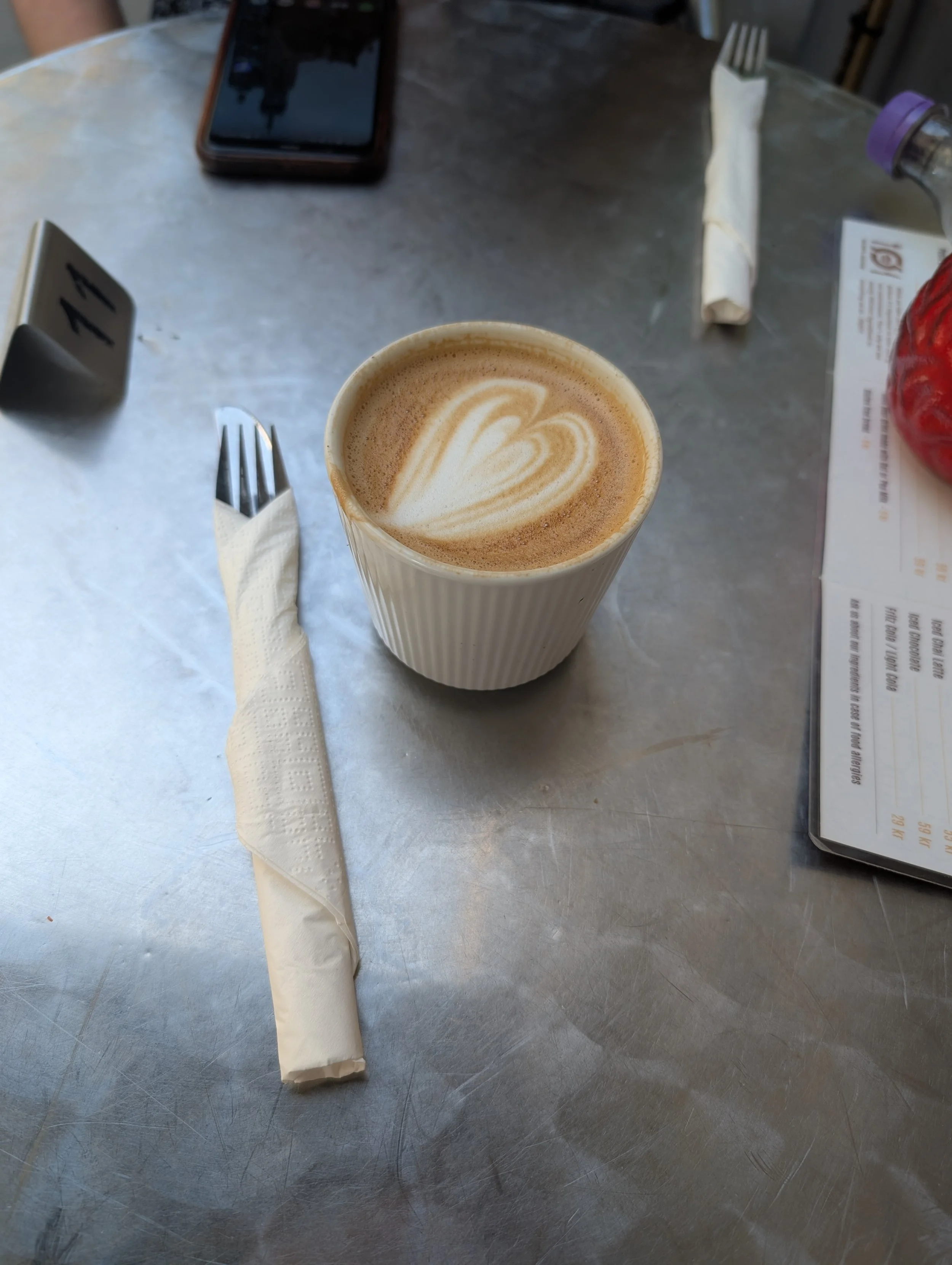 A table with a latte in a to-go cup, a fork wrapped in a napkin, a menu, a smartphone, an empty water bottle, and a wrapped straw.