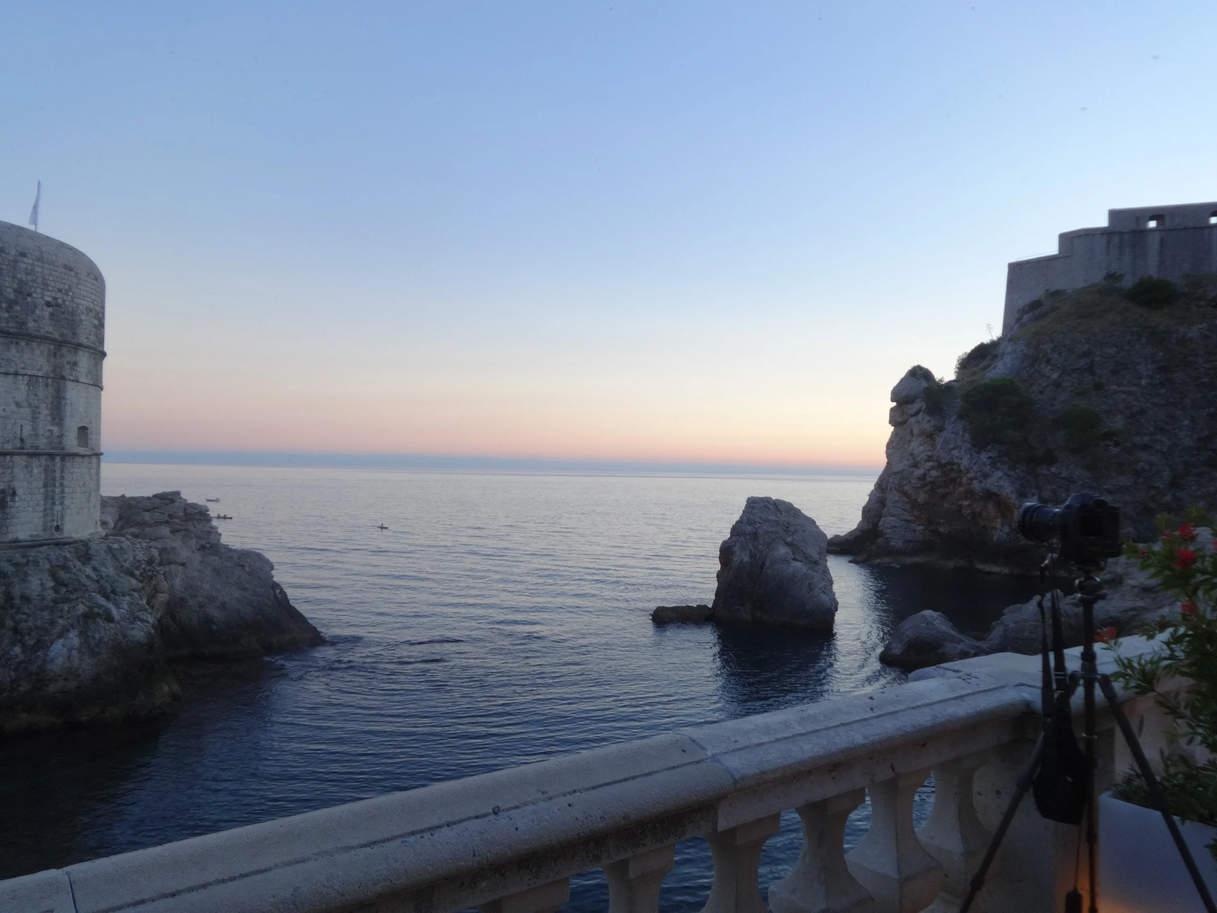 View of a coastal fortress or castle on the rocky shore, with the sea and sky at sunset or sunrise, and a telescope on a railing in the foreground.