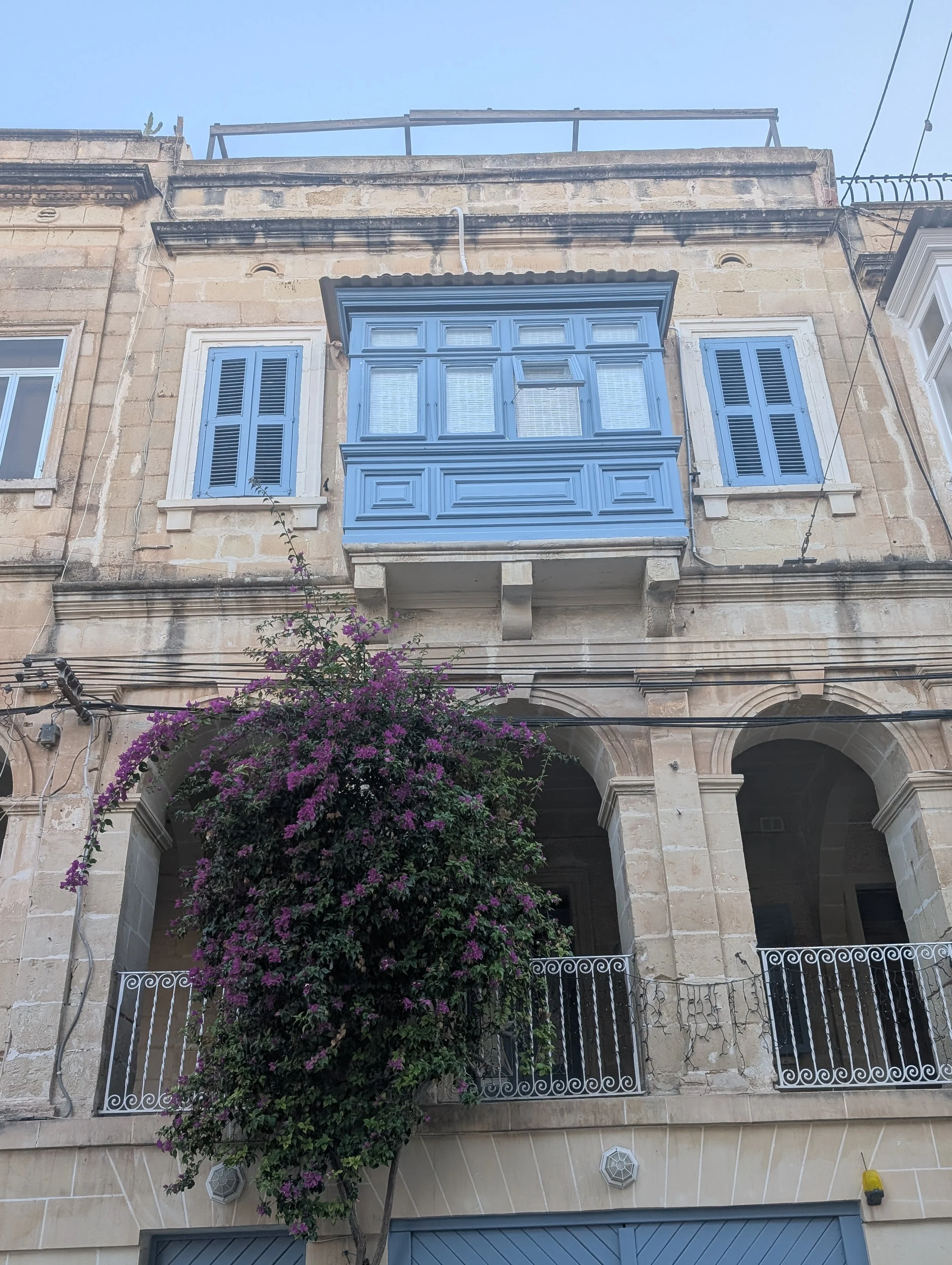A beige stone building with a blue wooden balcony and blue shutters on the windows. There is a purple flowering bush below the balcony and a wrought iron fence in front.