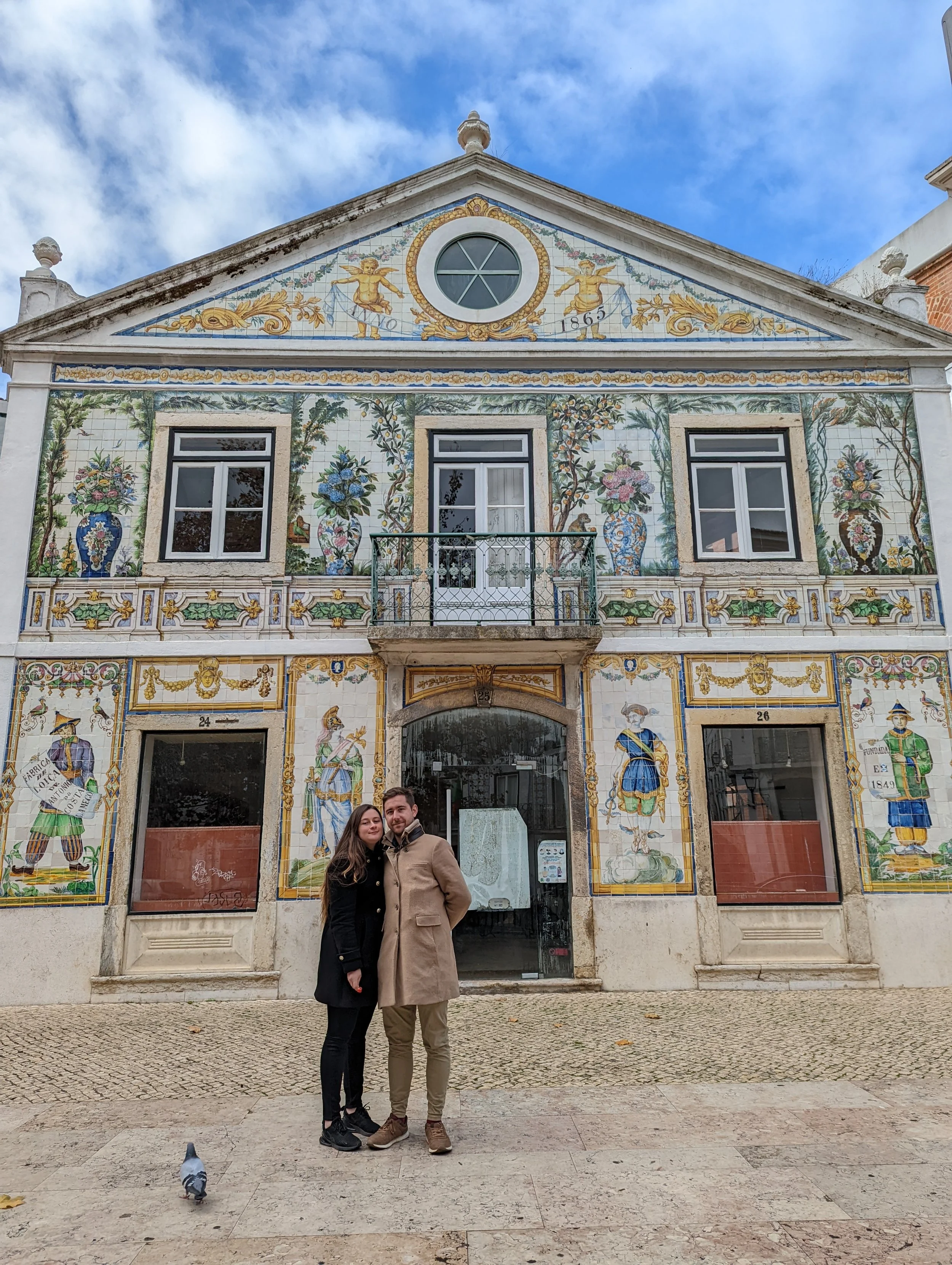 A couple standing in front of a historic tile-decorated building on a cloudy day. The woman is wearing a black coat and the man a beige coat. There is a pigeon on the ground near their feet.